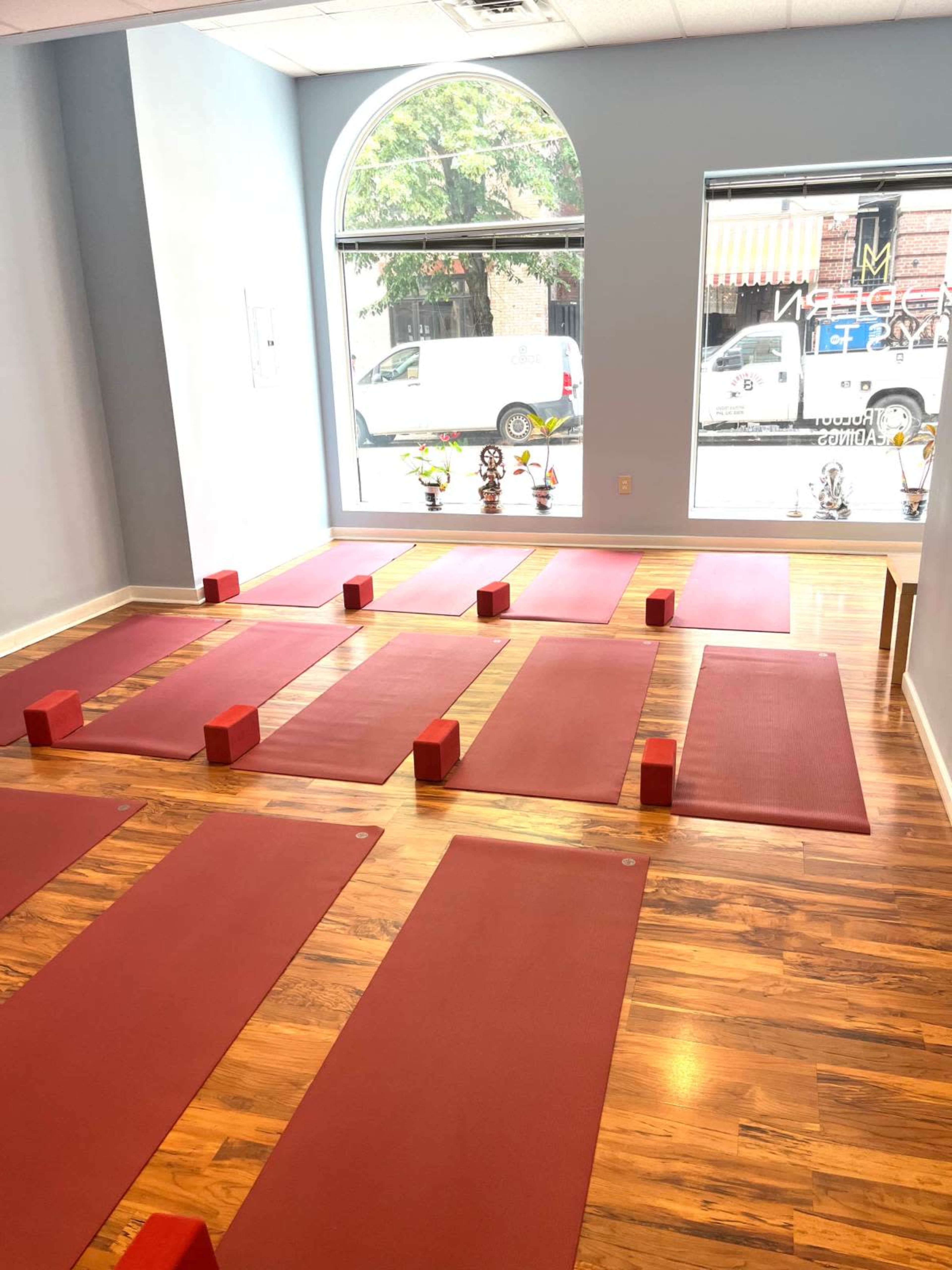 The image shows a yoga studio featuring red mats arranged neatly on a wooden floor, with blocks placed at the head of each mat and a large window allowing natural light to enter.