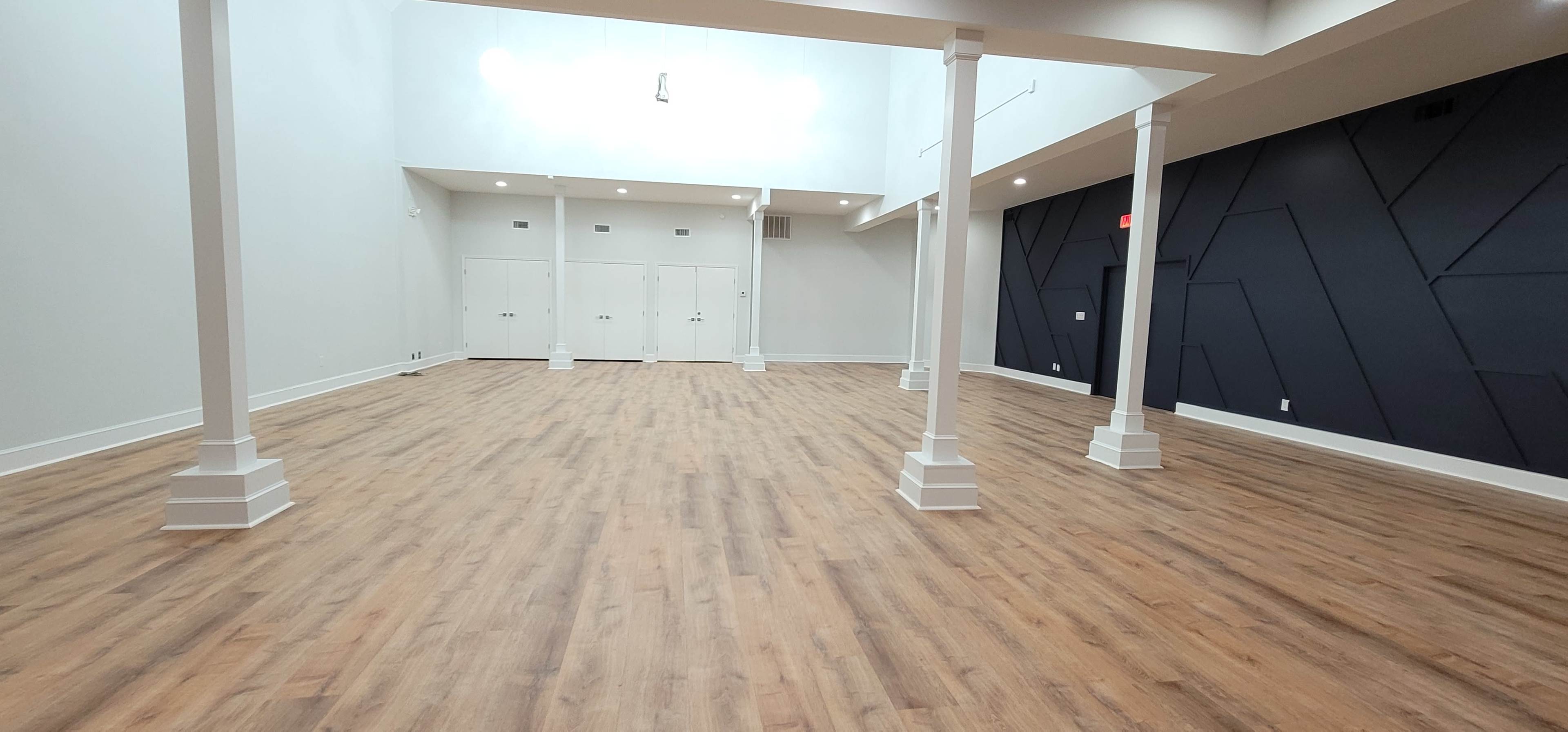 A spacious, empty room with wooden flooring, several white columns, and a dark accent wall.