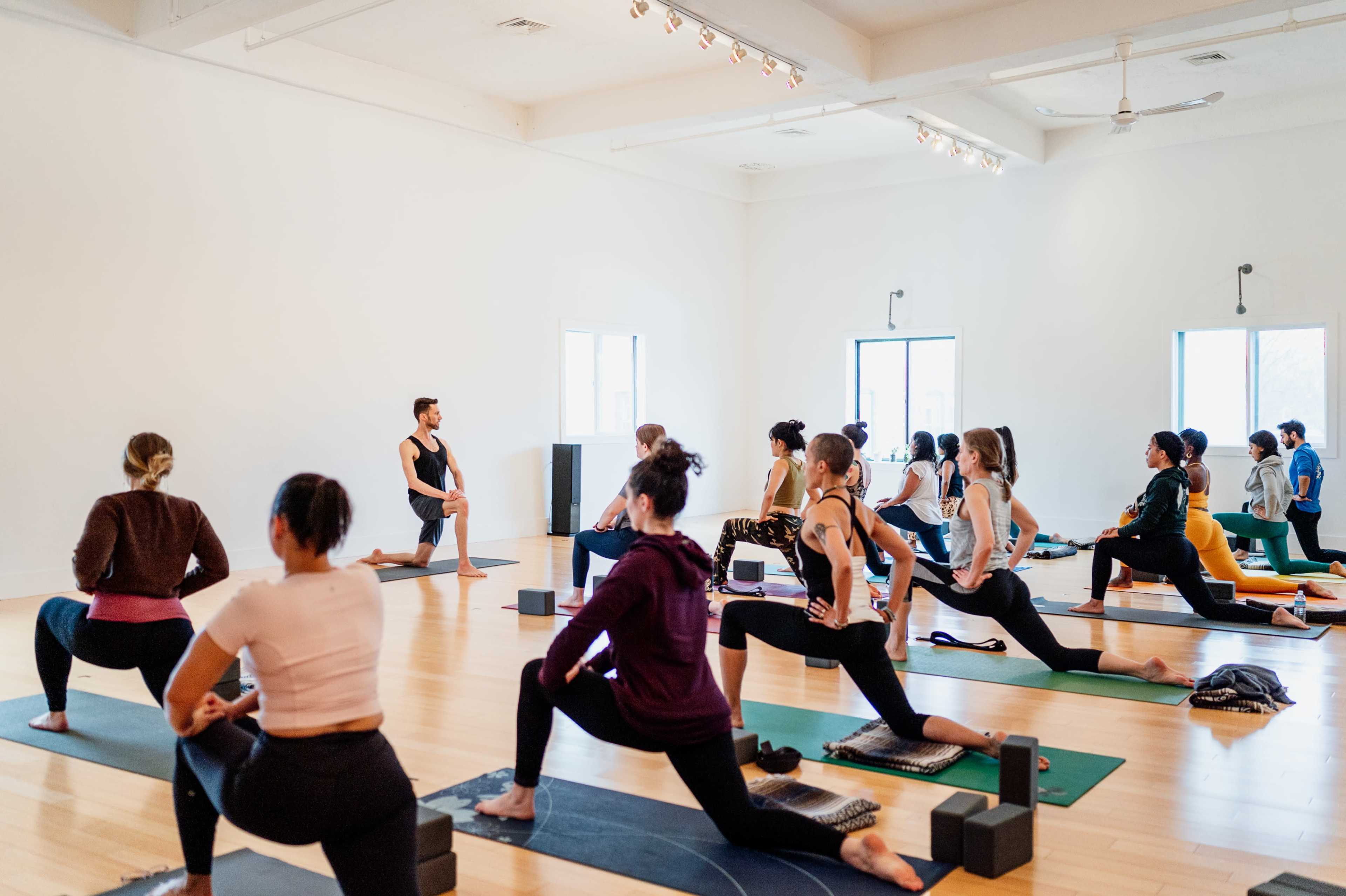 A yoga instructor guides a class of participants in a spacious studio, with students practicing lunges on their mats.