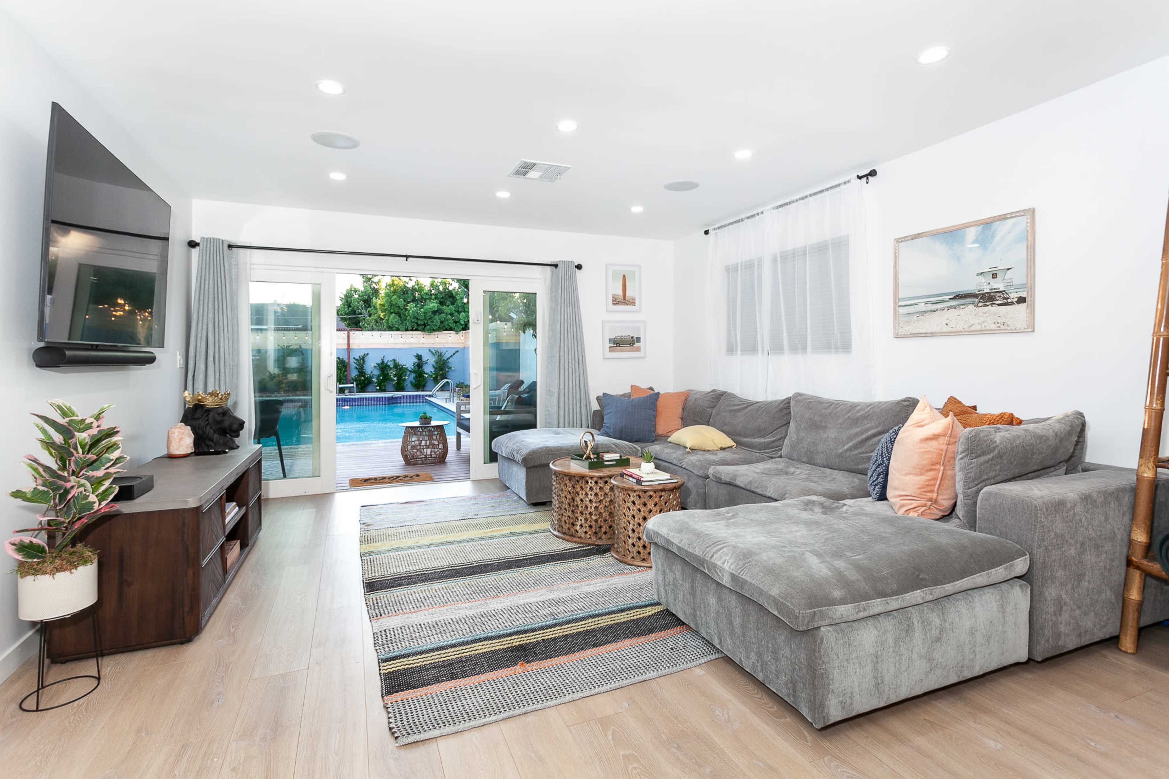 The living room features a large gray sectional sofa, a wooden TV stand, and sliding glass doors that open to a pool area, complemented by a striped area rug and decorative accents.