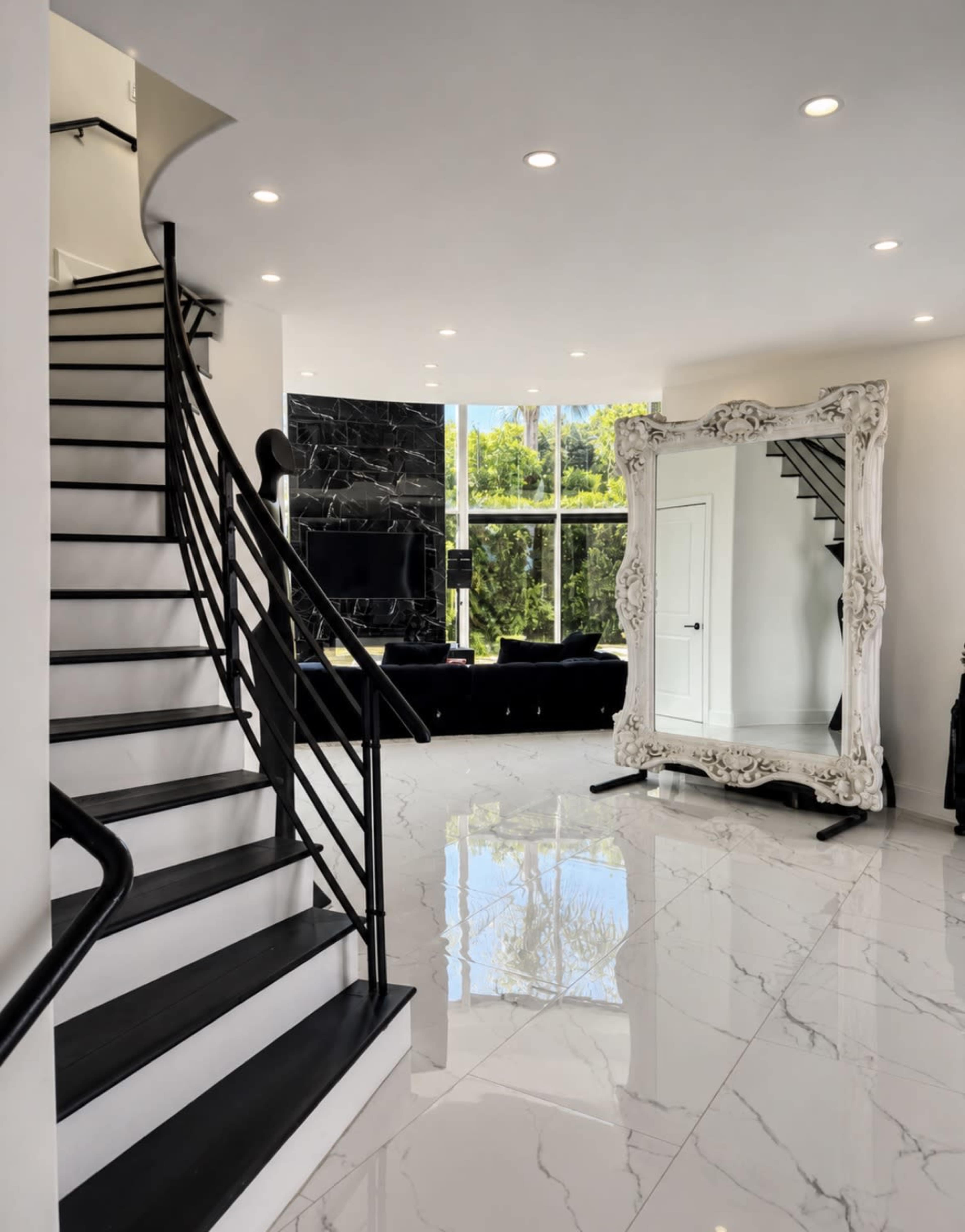 The image shows a modern foyer with a spiral staircase, large decorative mirror, and a living area featuring a black couch and large windows.