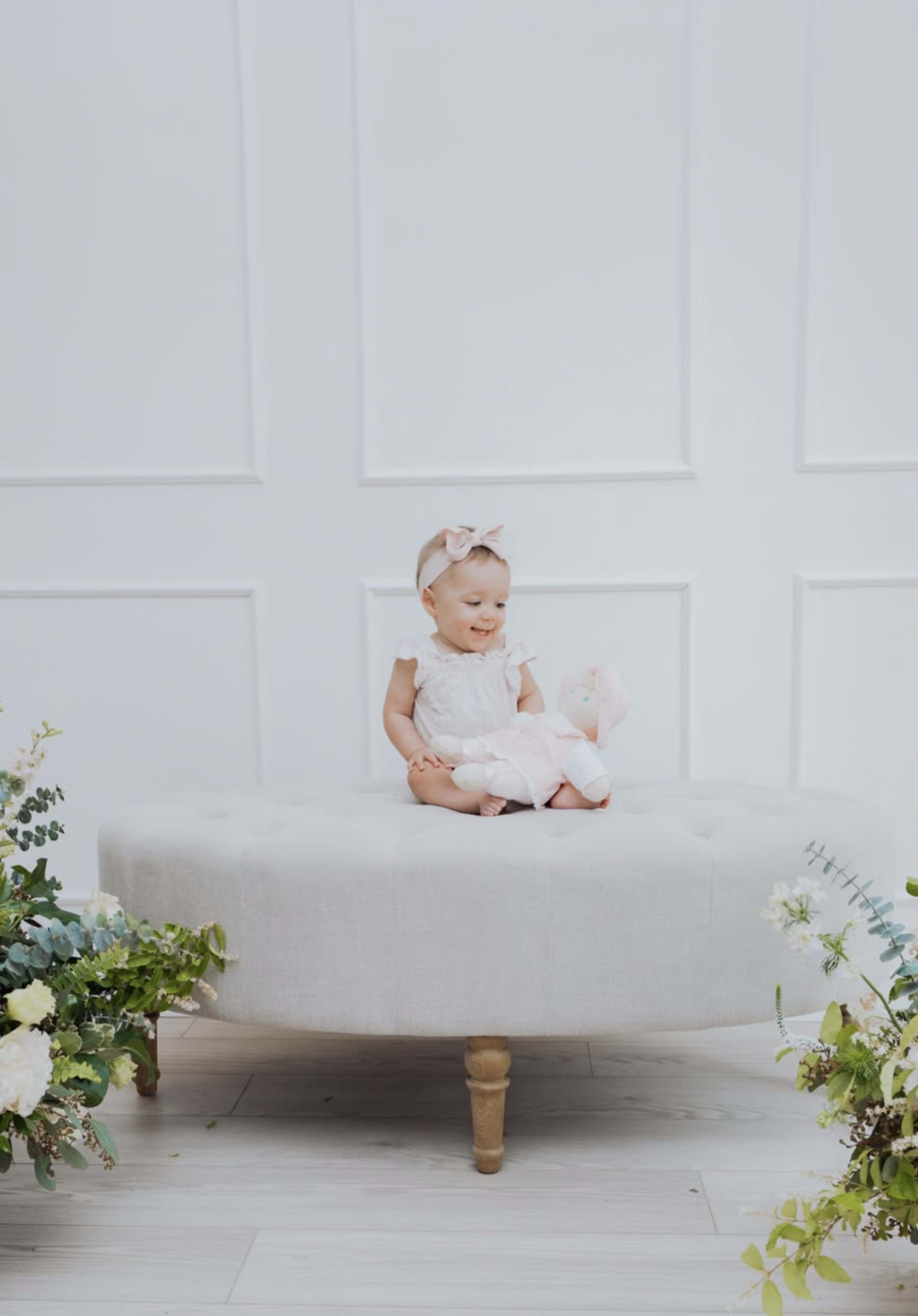 A child sits on a light-colored tufted ottoman holding a stuffed animal, framed by greenery and flowers.