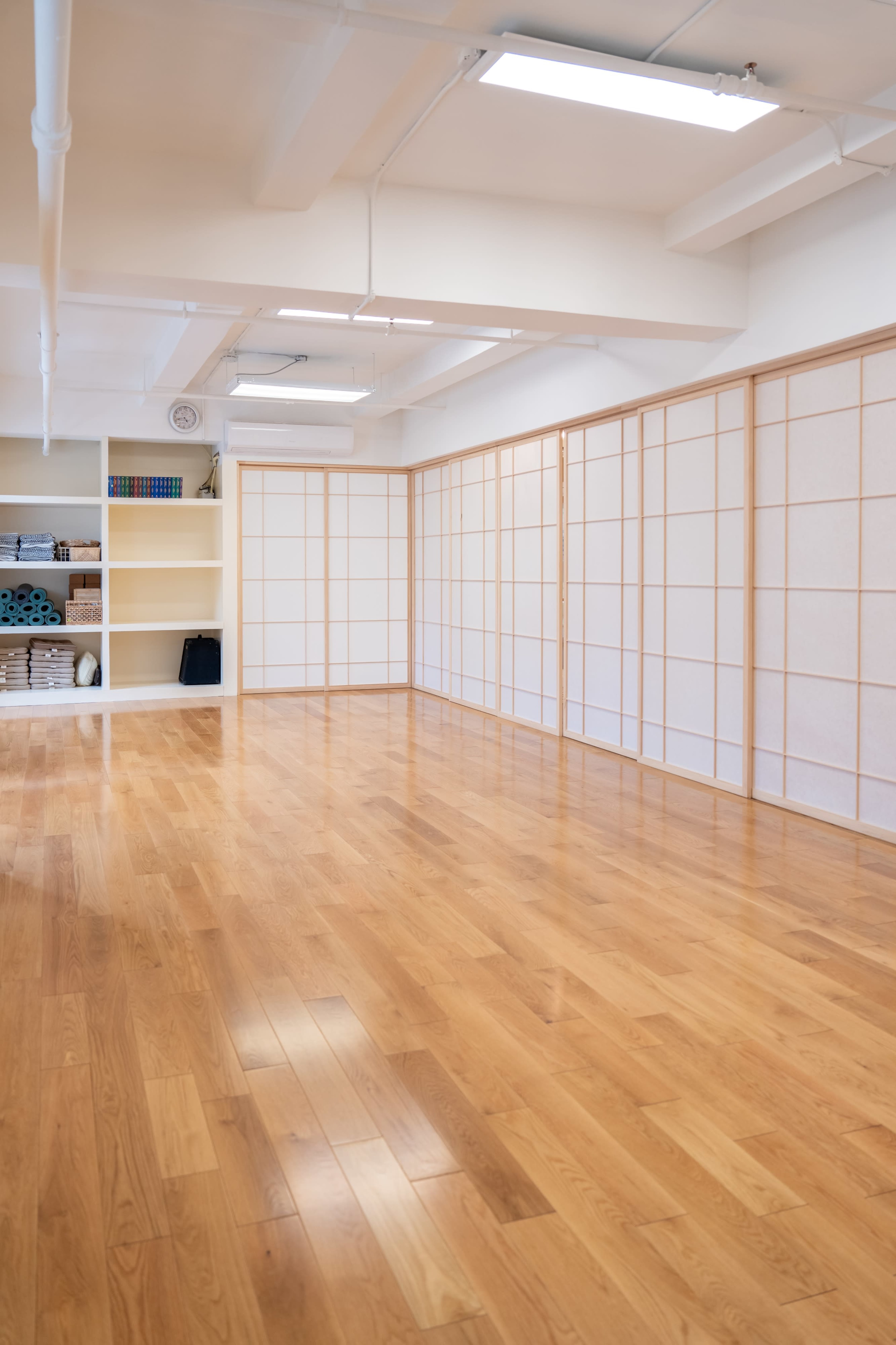 The image shows a spacious, well-lit studio with wooden floors and minimalist decor, featuring a shelving unit and sliding paper panels.