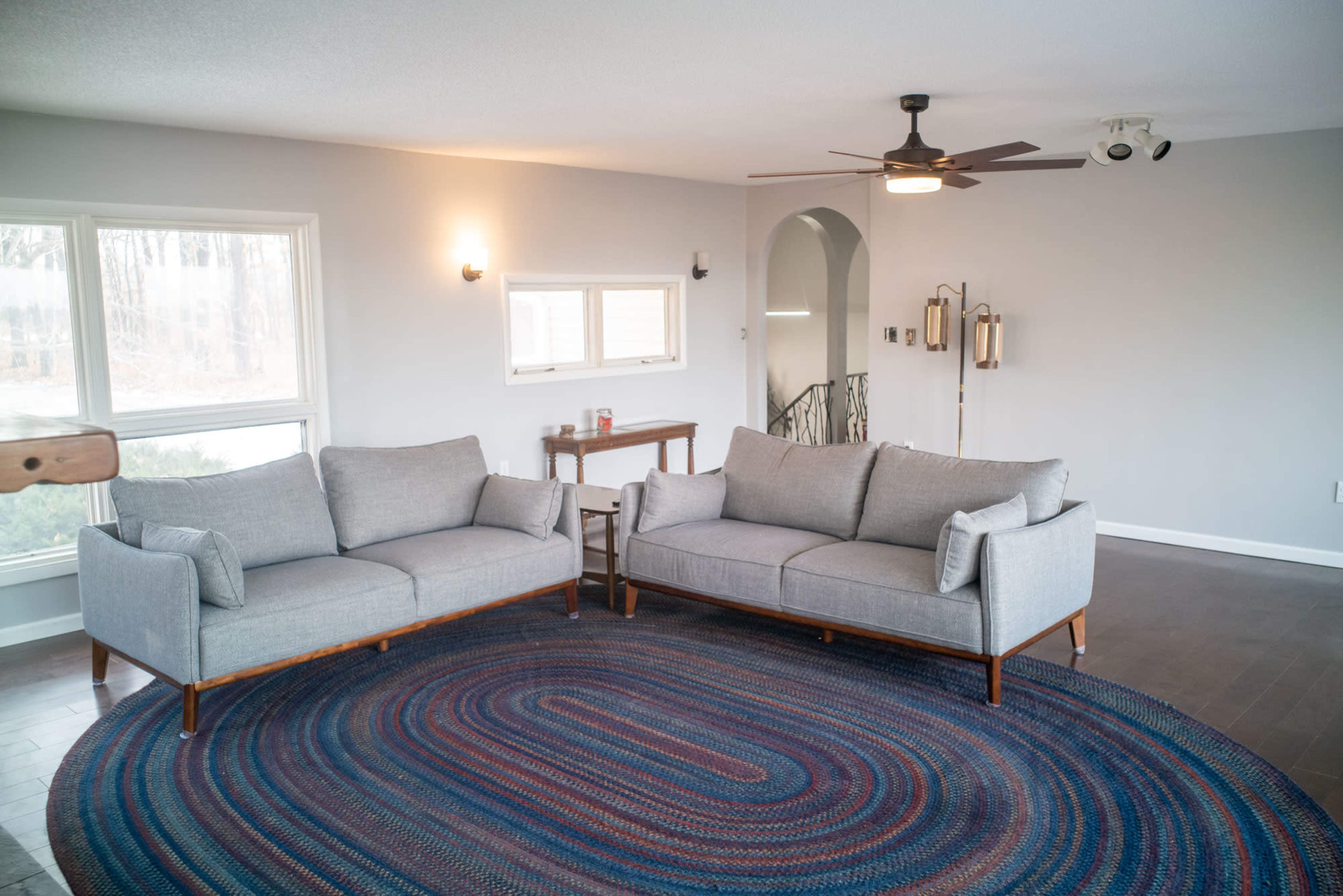 A spacious living room features two gray sofas facing each other on a circular area rug, with large windows providing natural light.