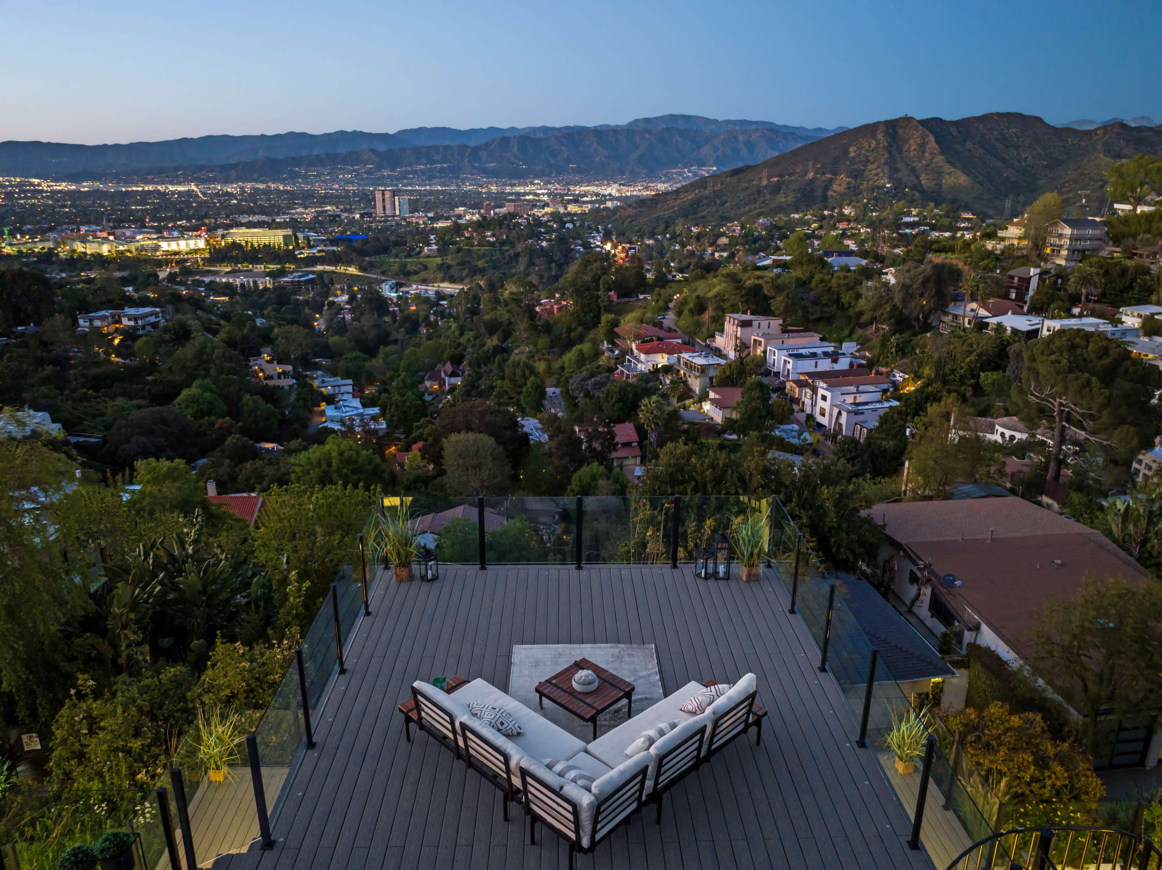 A spacious outdoor terrace with a sectional sofa overlooks a valley filled with homes and mountains at dusk.
