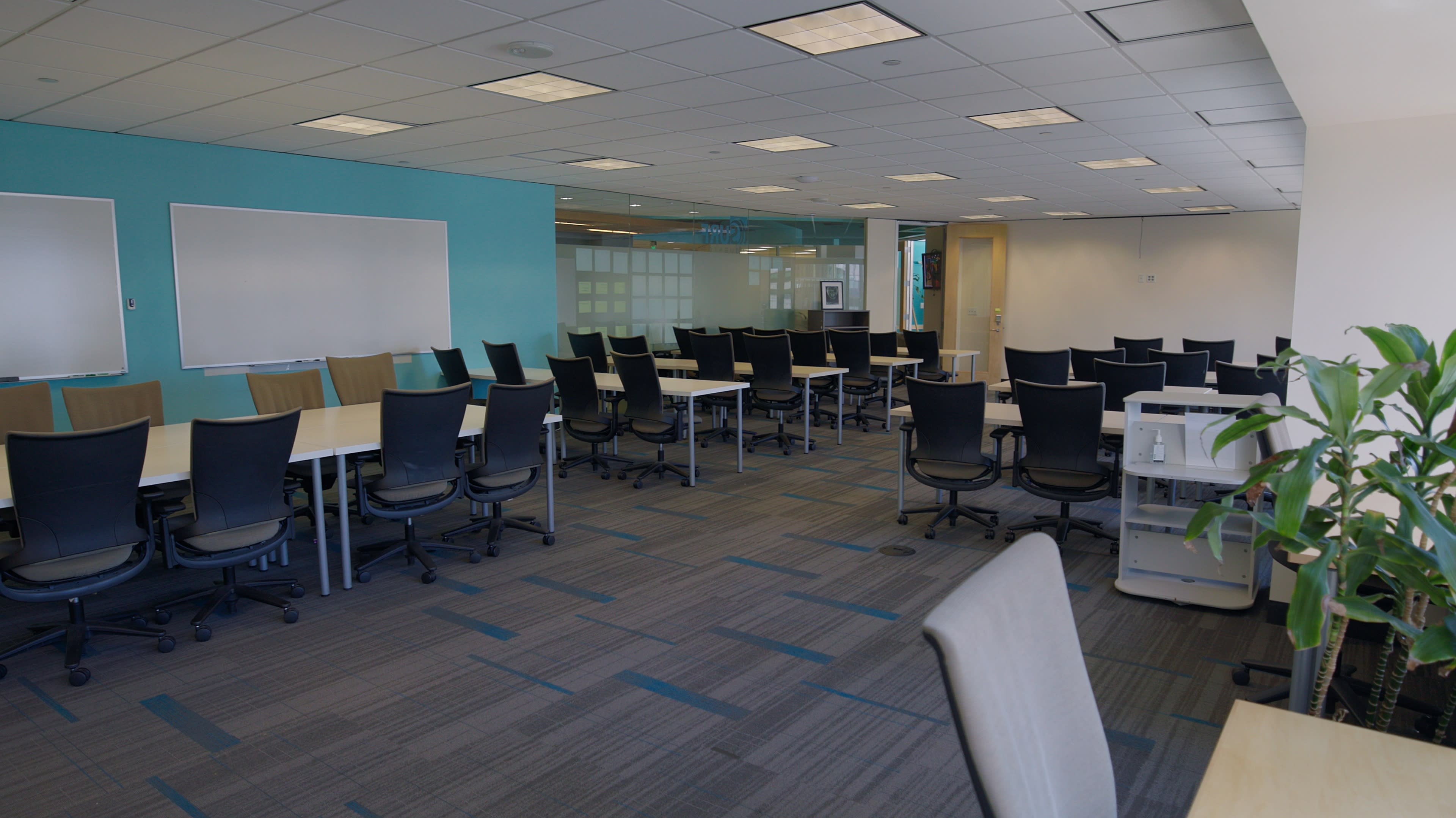 The image shows a modern office space with multiple rows of tables and ergonomic chairs arranged neatly, and a green accent wall in the background.
