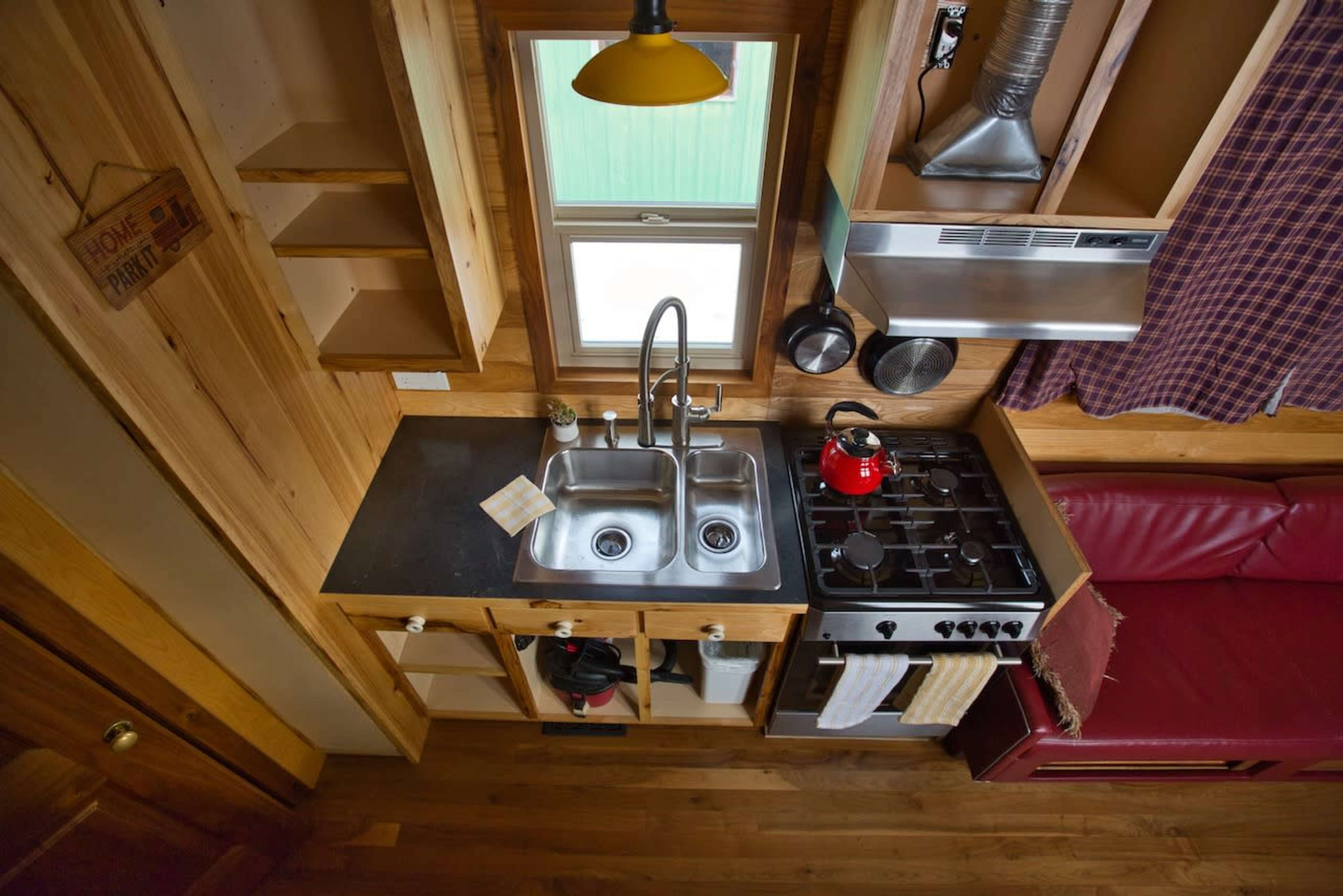 A compact kitchen with a double sink, a gas stove, and wooden cabinetry, featuring a red kettle on the stove and a small dining area nearby.
