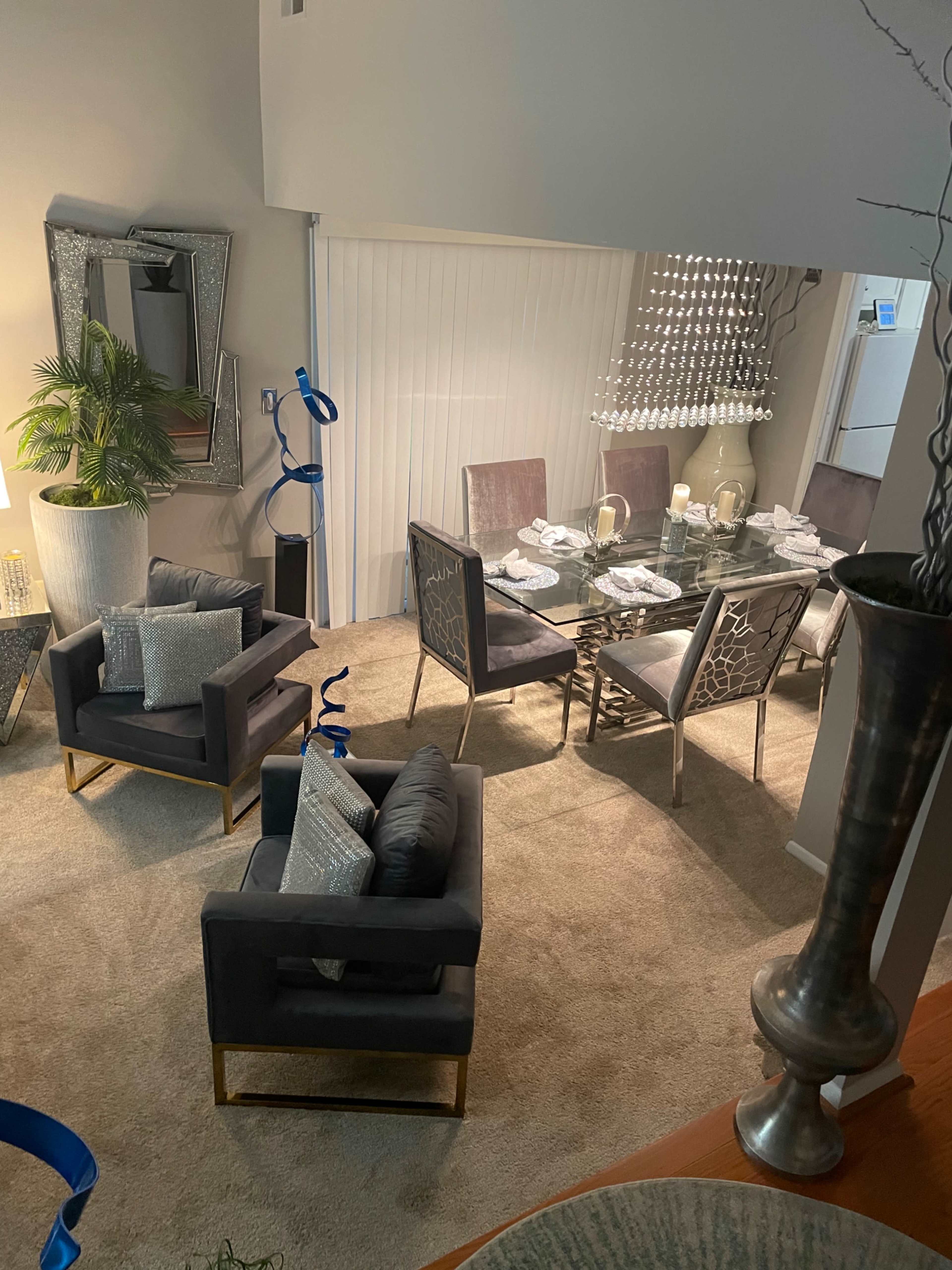 A modern dining area featuring a glass table surrounded by stylish chairs, with two armchairs positioned nearby and large windows draped with vertical blinds.