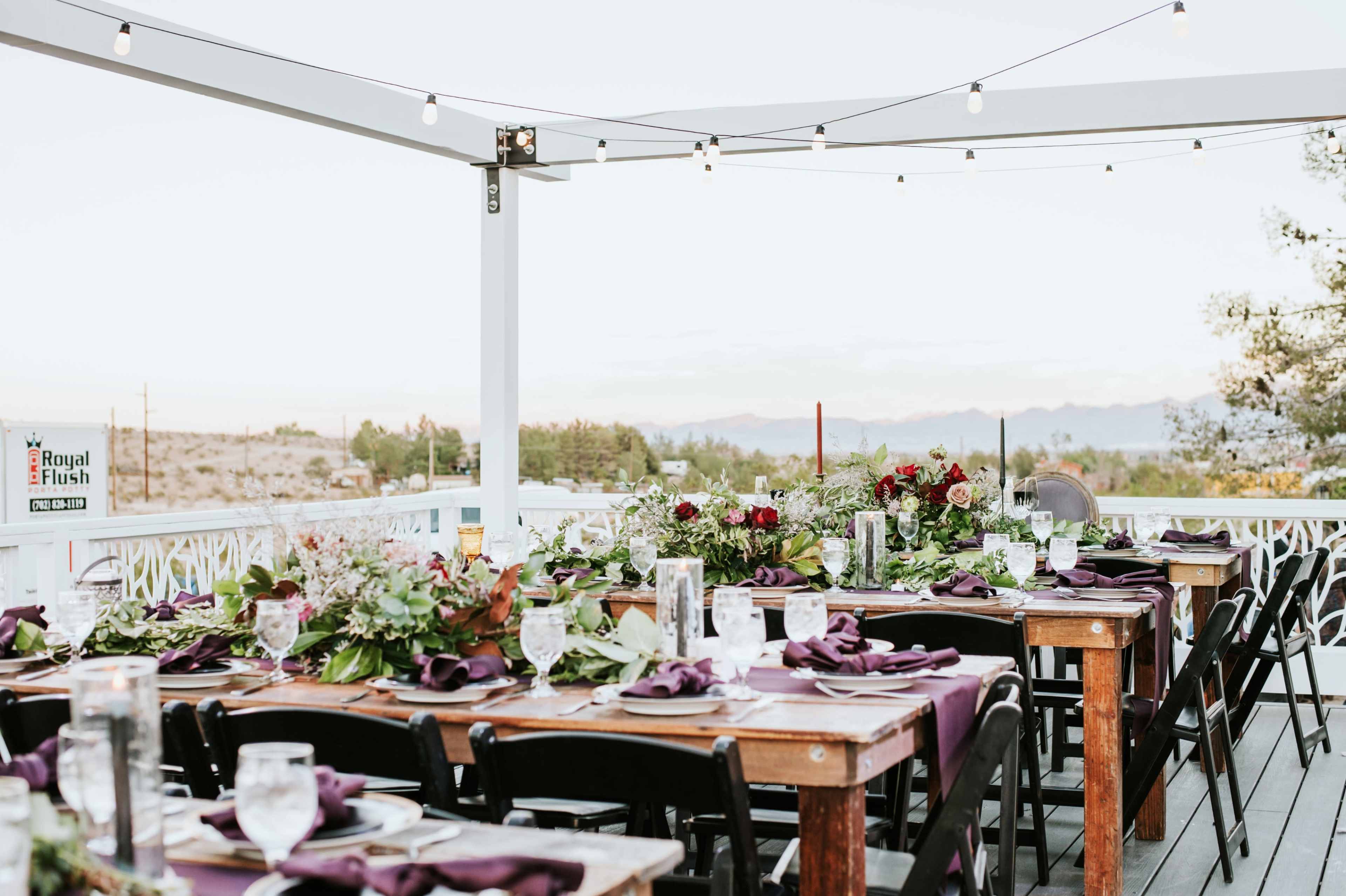 A long dining table is set on a patio with greenery and floral arrangements, surrounded by mountains under a string of lights.