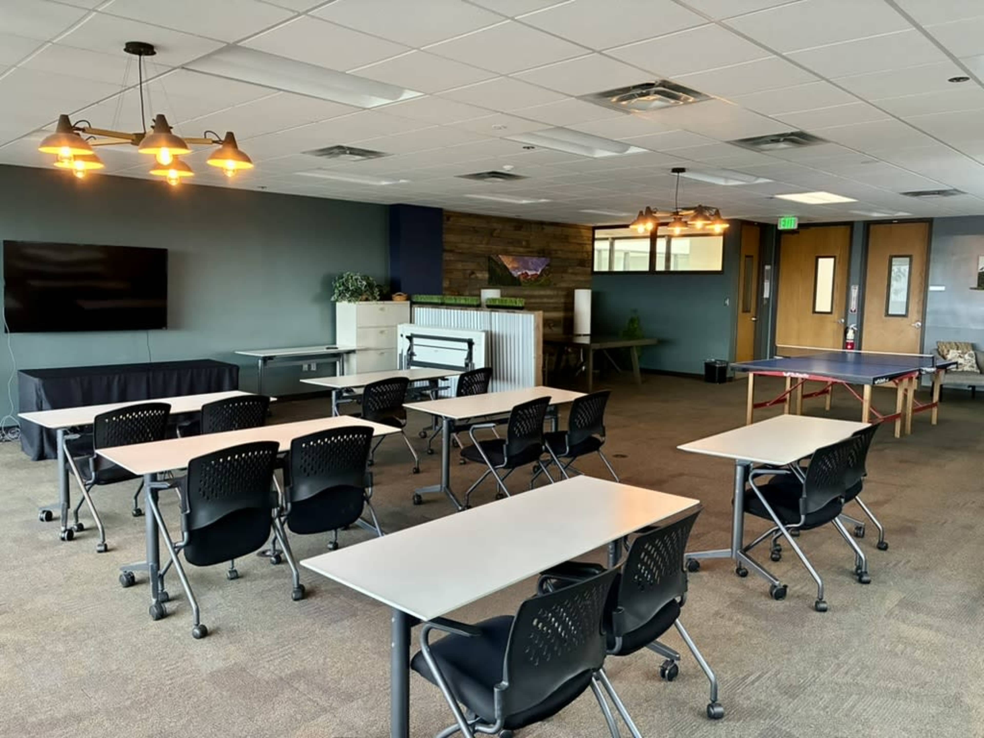 The image shows a spacious room with several tables and chairs arranged for meetings, a large screen on one wall, and recreational equipment in the background.
