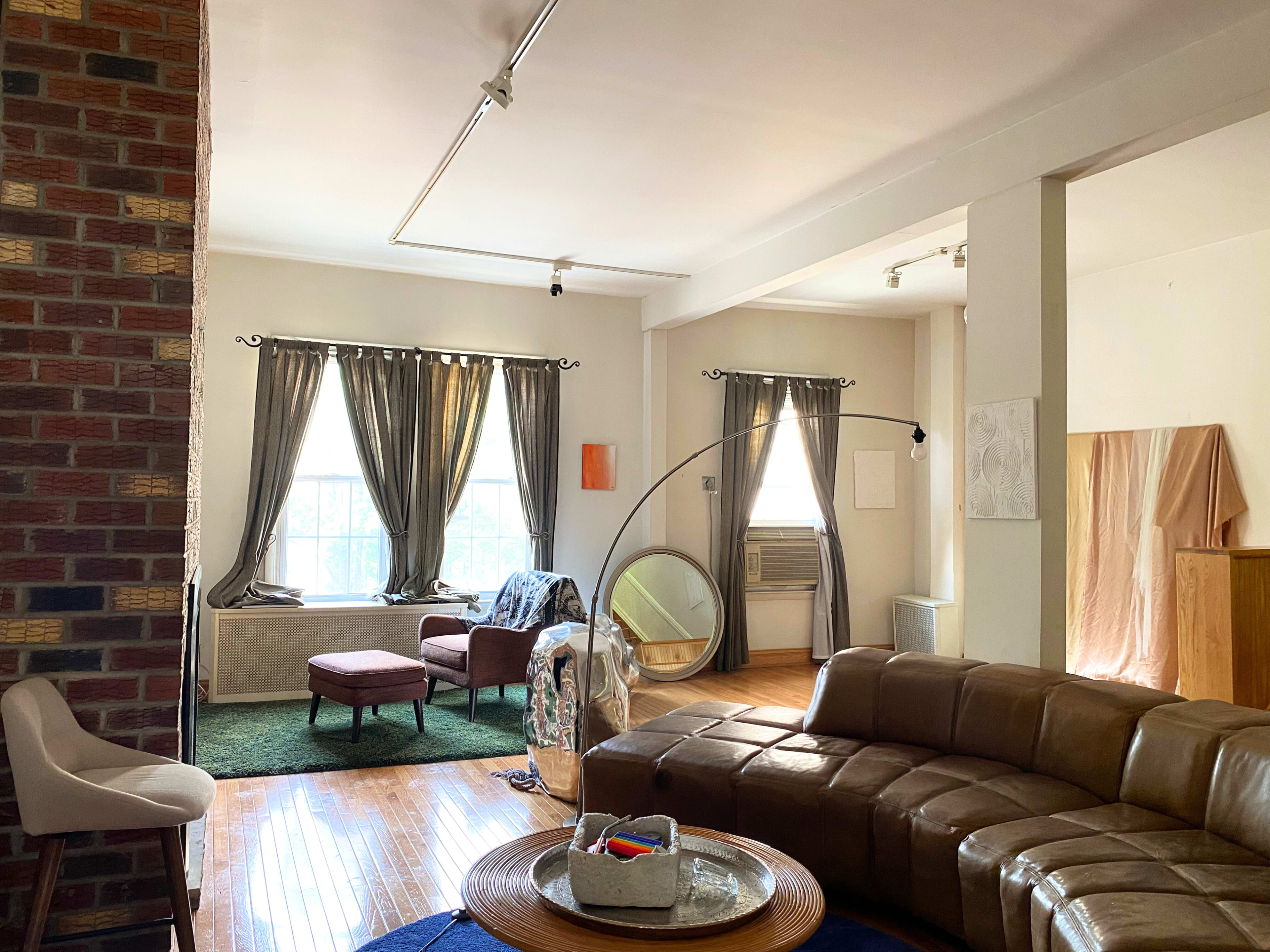 A spacious living room with large windows, a mix of furniture including a curved couch and a chair, and a decorative mirror placed near a green rug.