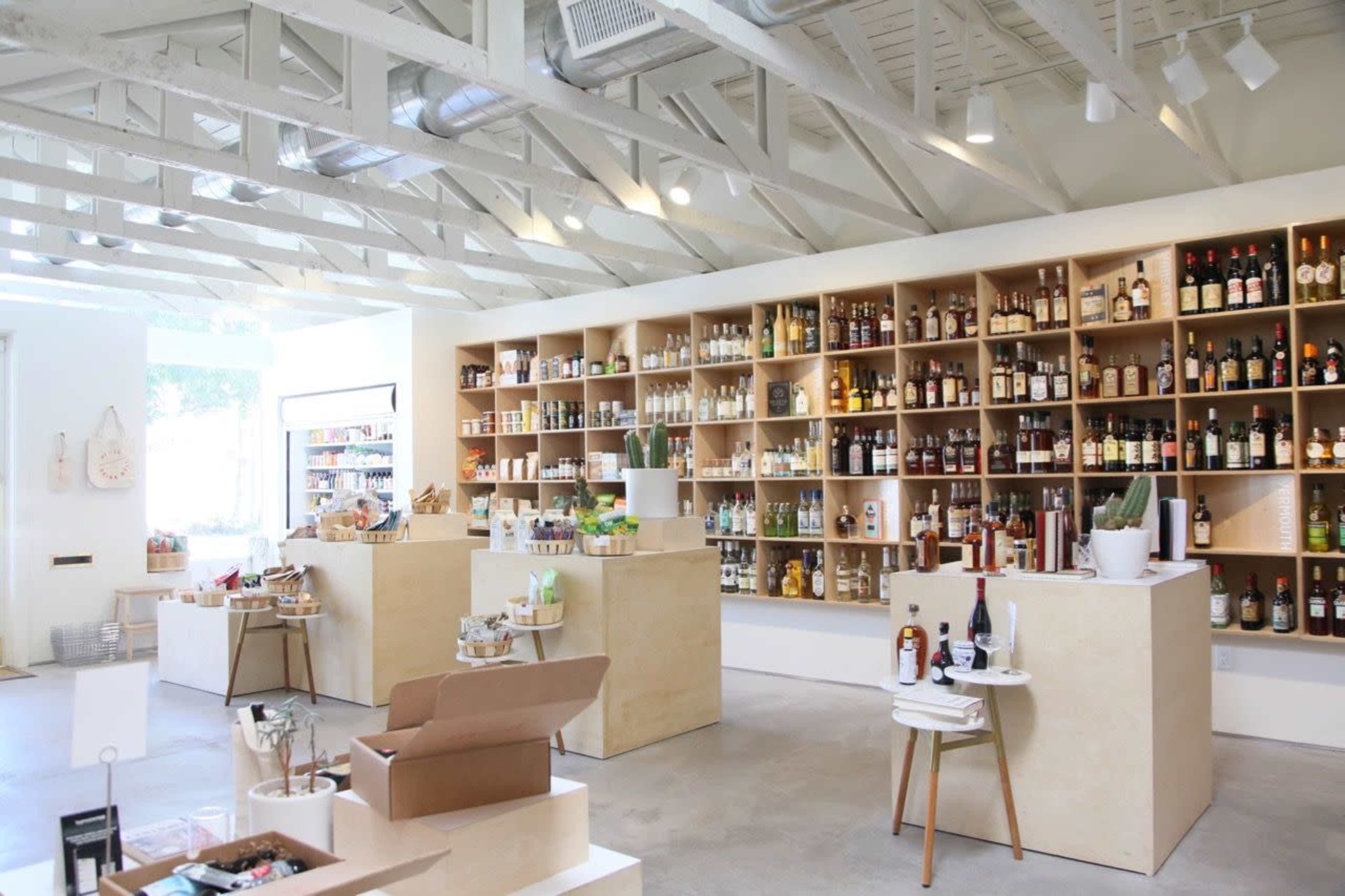 The image shows a modern retail space with wooden shelving filled with various bottles and food items, and minimalist display tables throughout the room.