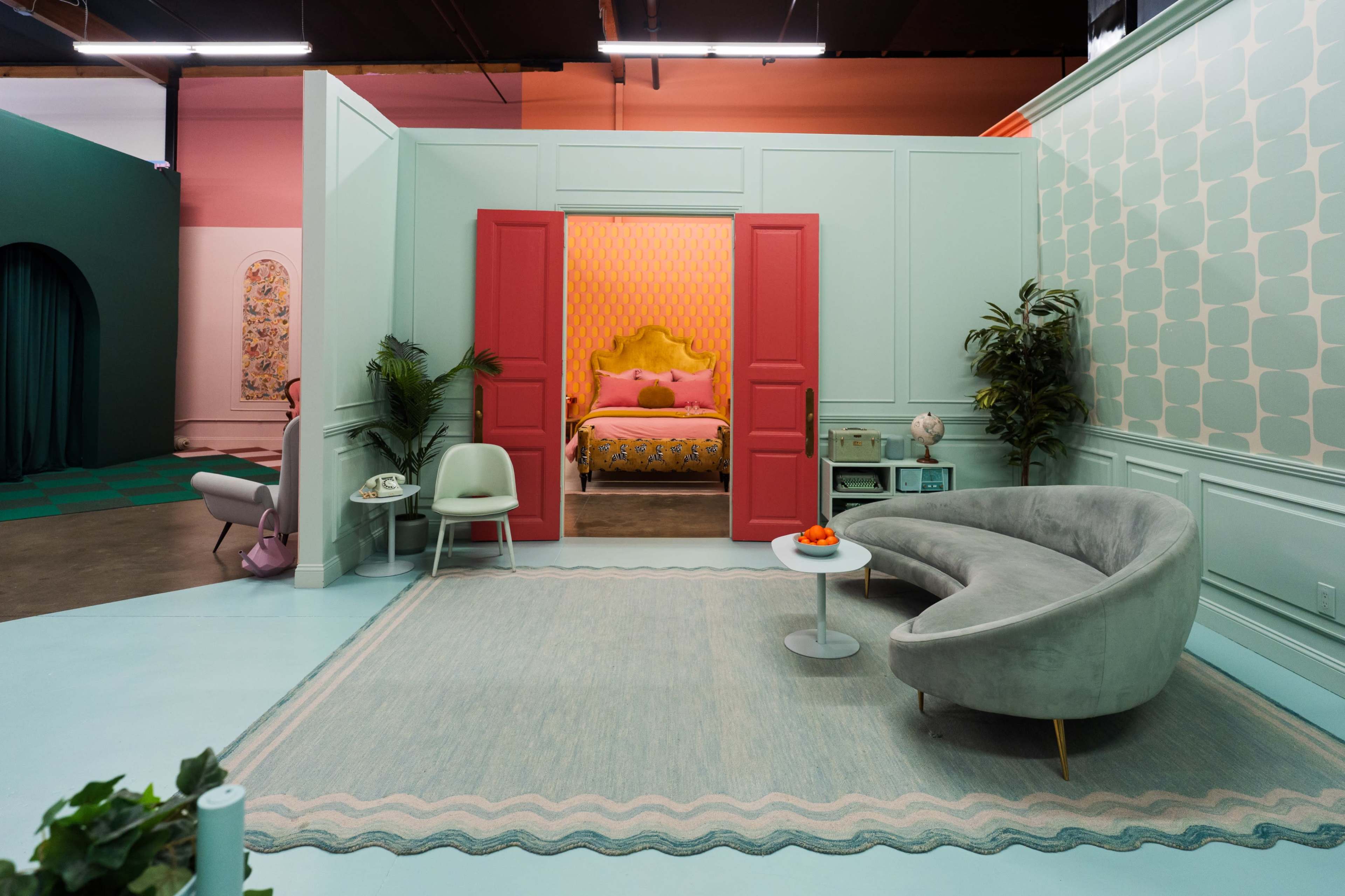 The image shows a modern interior scene featuring a living area with a curved sofa, a circular table, and a doorway leading to a brightly colored bedroom.