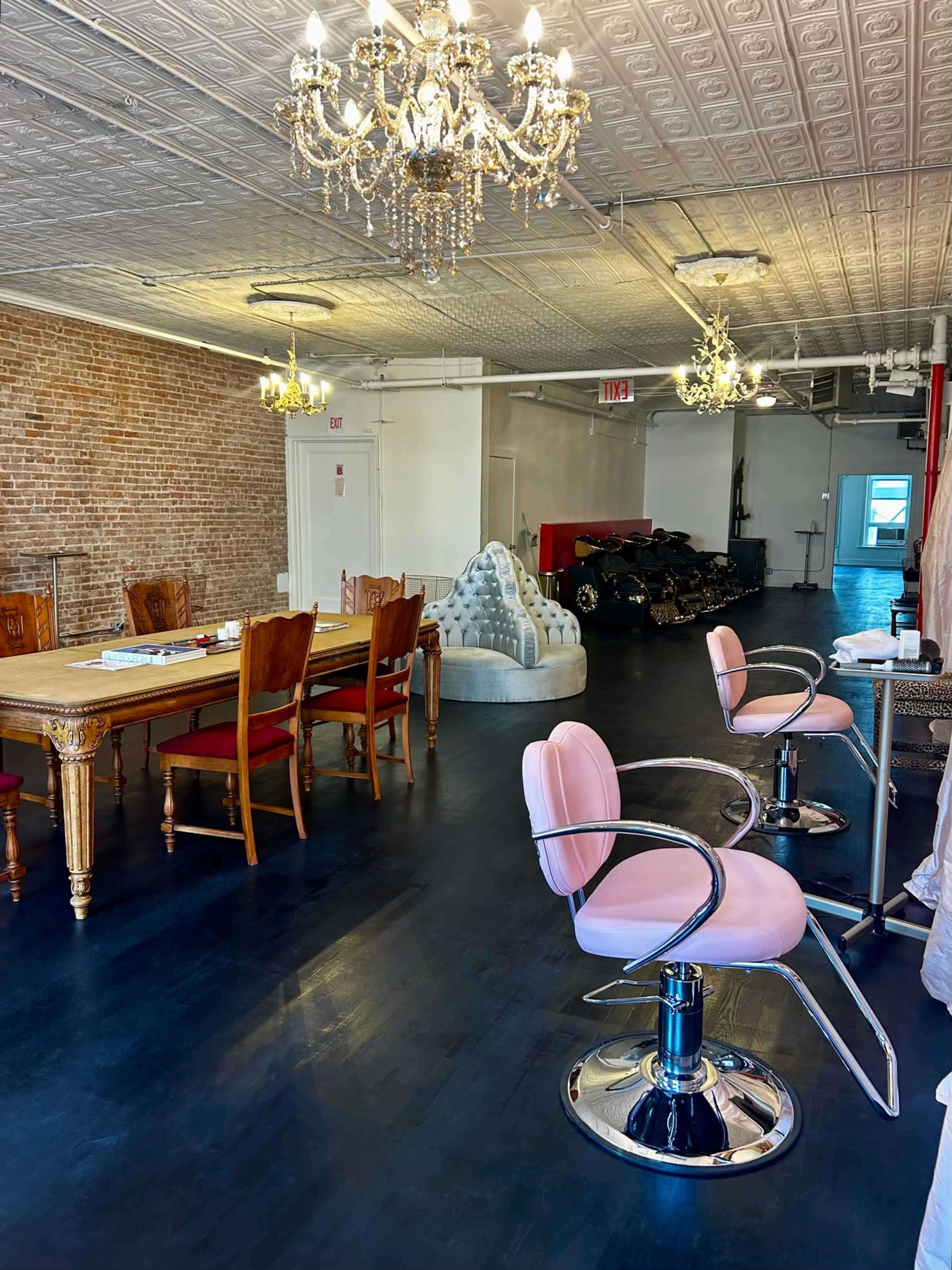 A spacious interior with a wooden dining table, pink salon chairs, and chandeliers, featuring exposed brick walls and a variety of seating arrangements.