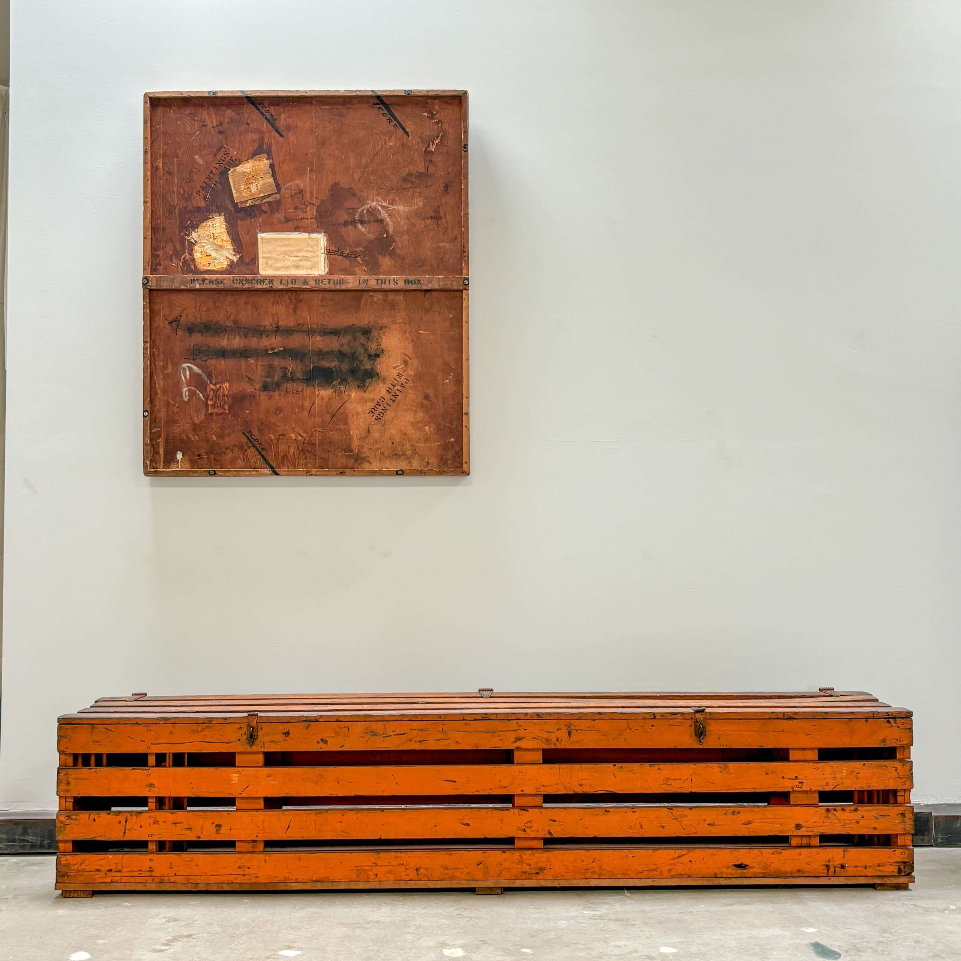 A wooden bench made of orange slats sits below a framed, rusted metal piece mounted on a white wall.