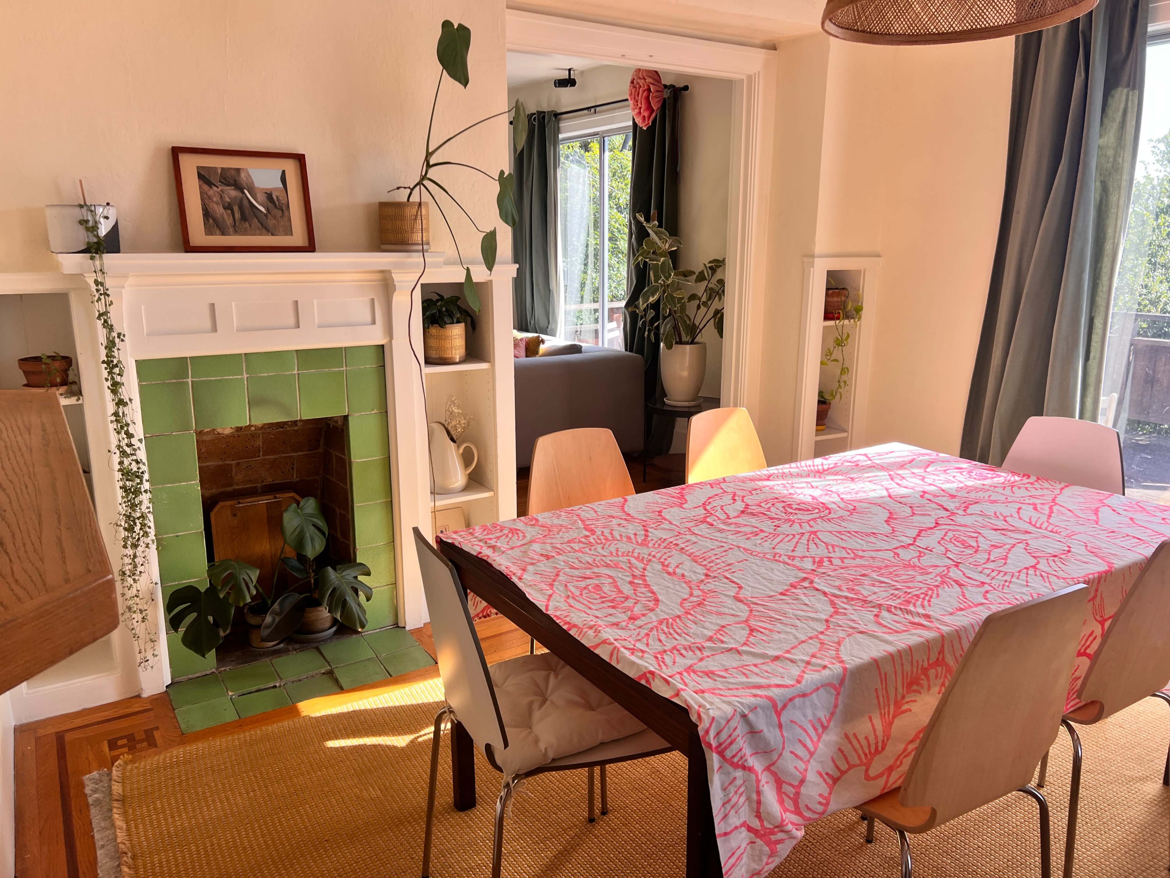 A dining area features a wooden table covered with a bright tablecloth, surrounded by chairs, alongside a green-tiled fireplace and large windows letting in natural light.