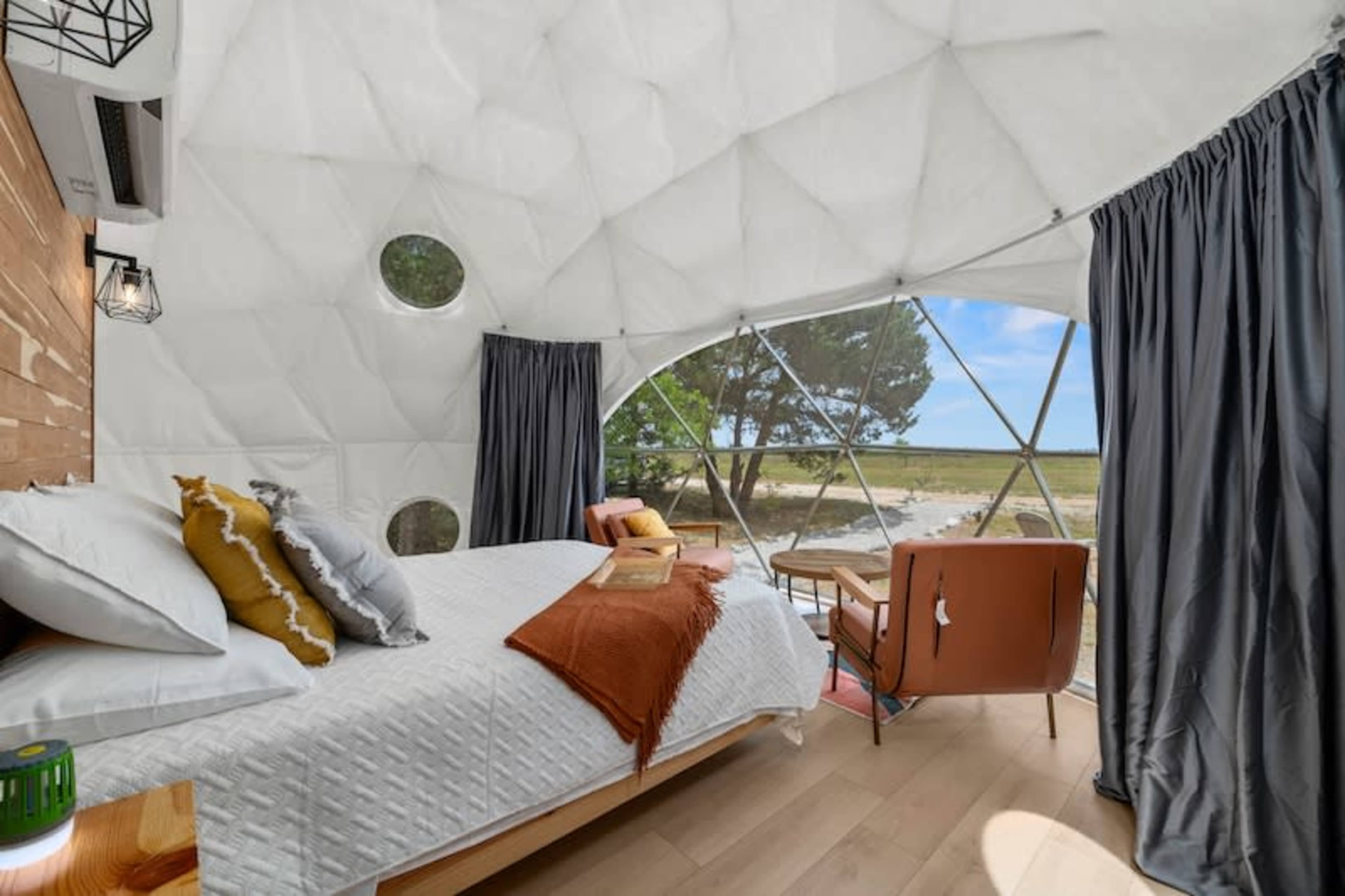 A modern geodesic dome interior featuring a bed, an armchair, and large windows overlooking a natural landscape.