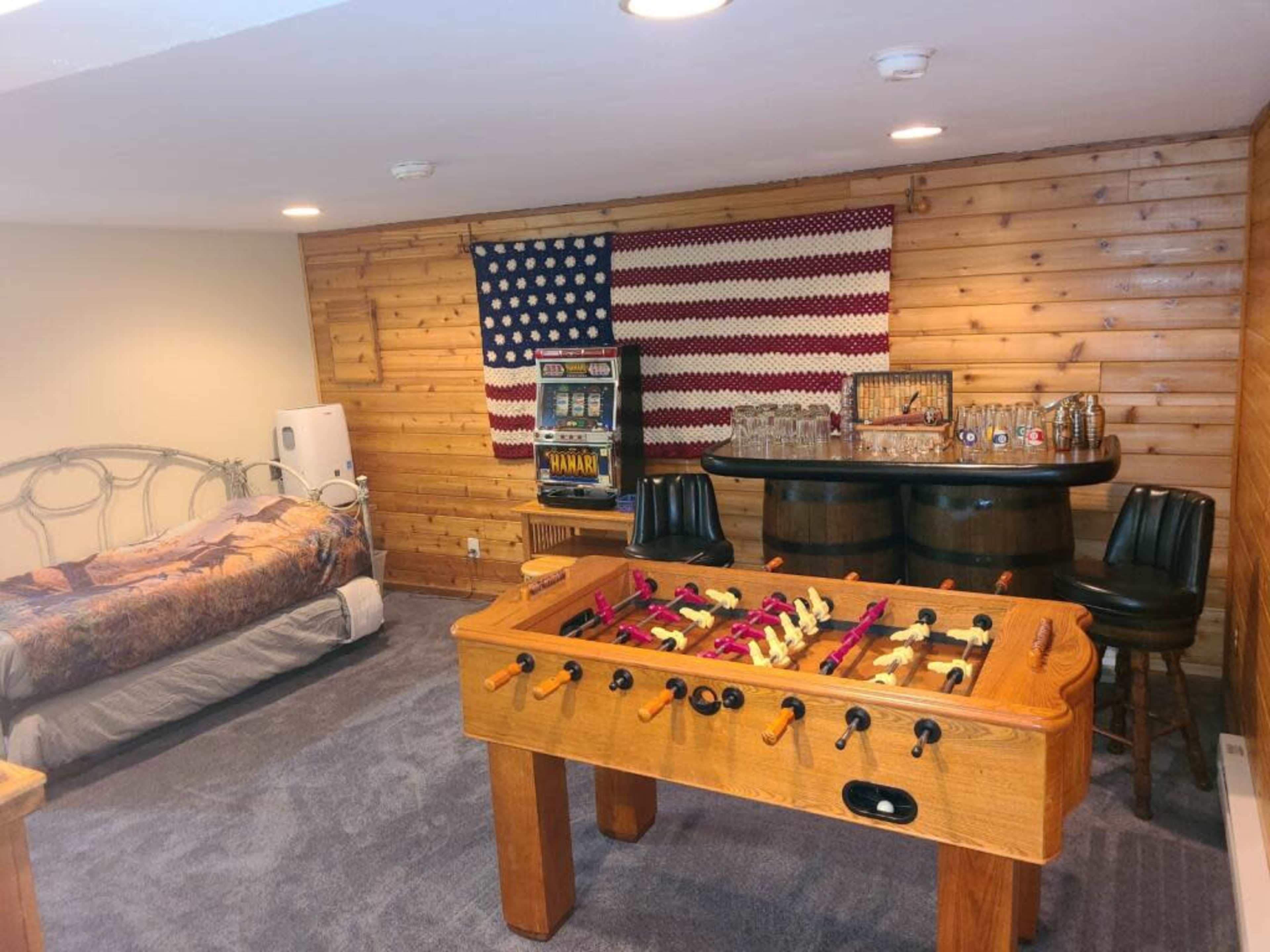 The image shows a basement room with a foosball table, a bed, a bar area with two stools, and a wall decorated with an American flag and wooden paneling.