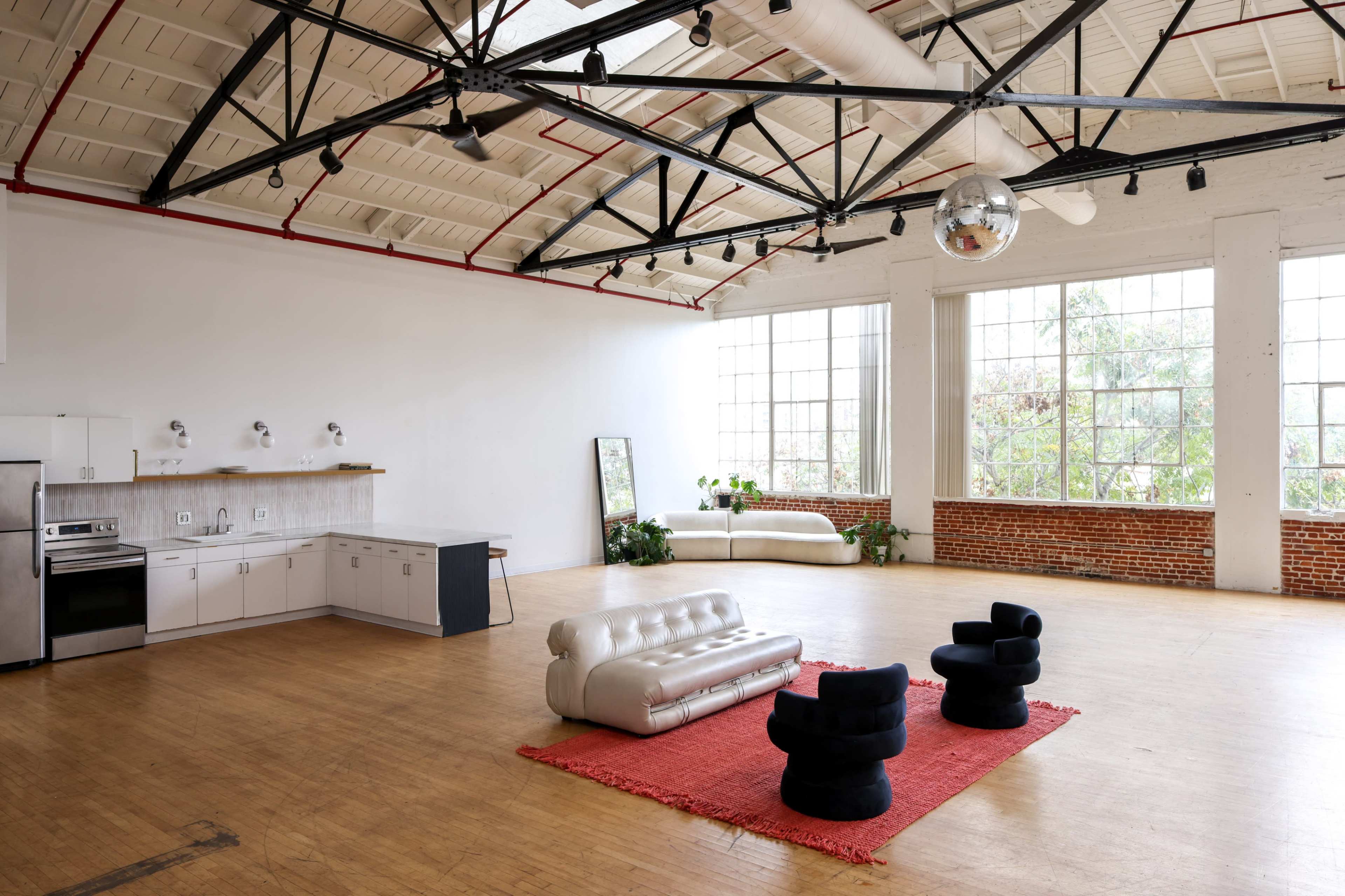 The image shows an industrial-style open space featuring a kitchen area, a silver sofa, two black chairs, and a large window allowing natural light to enter.