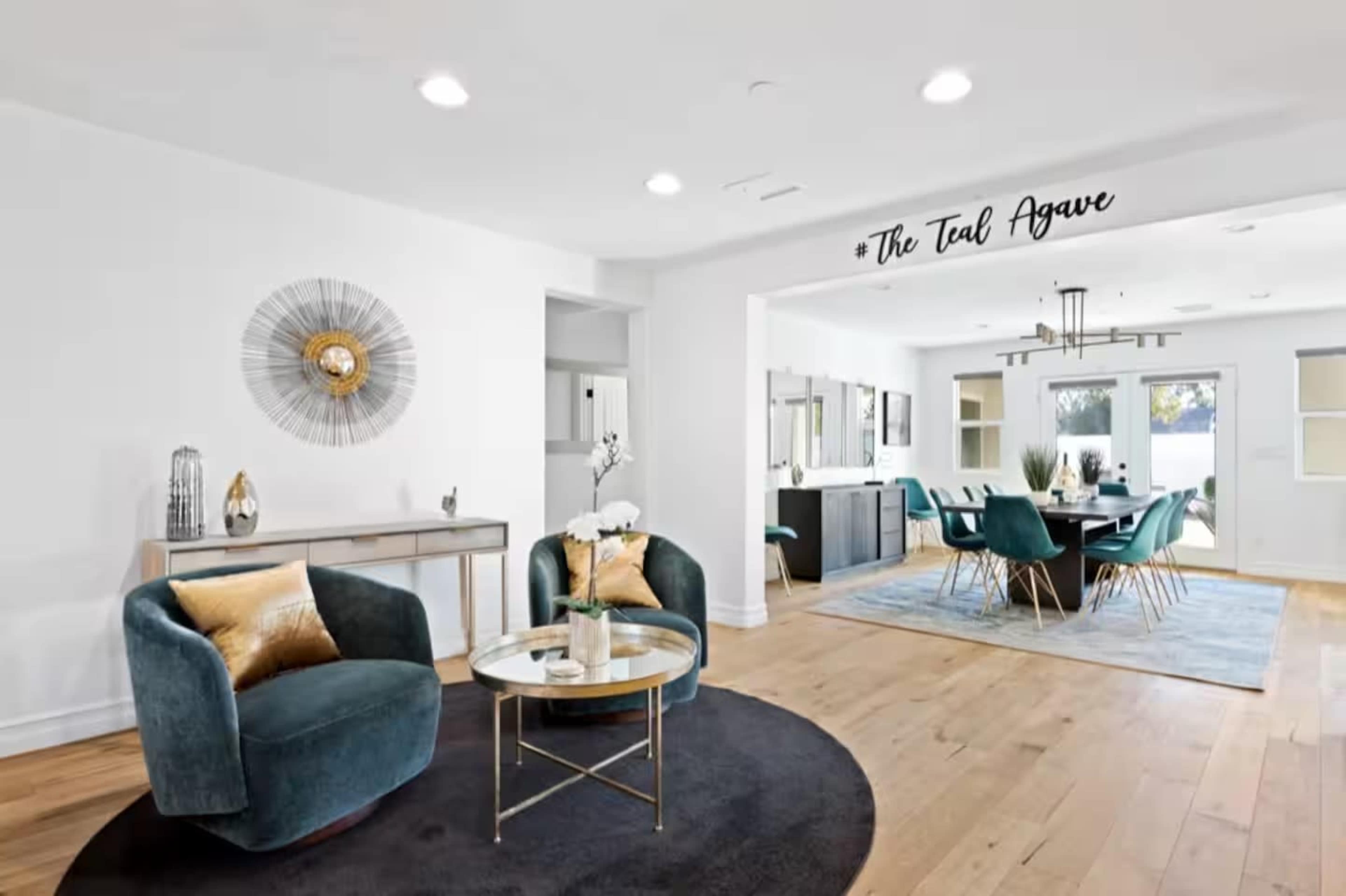 The image shows a modern living room featuring two teal armchairs and a round black rug, with a dining area visible in the background that includes a large table and green chairs.