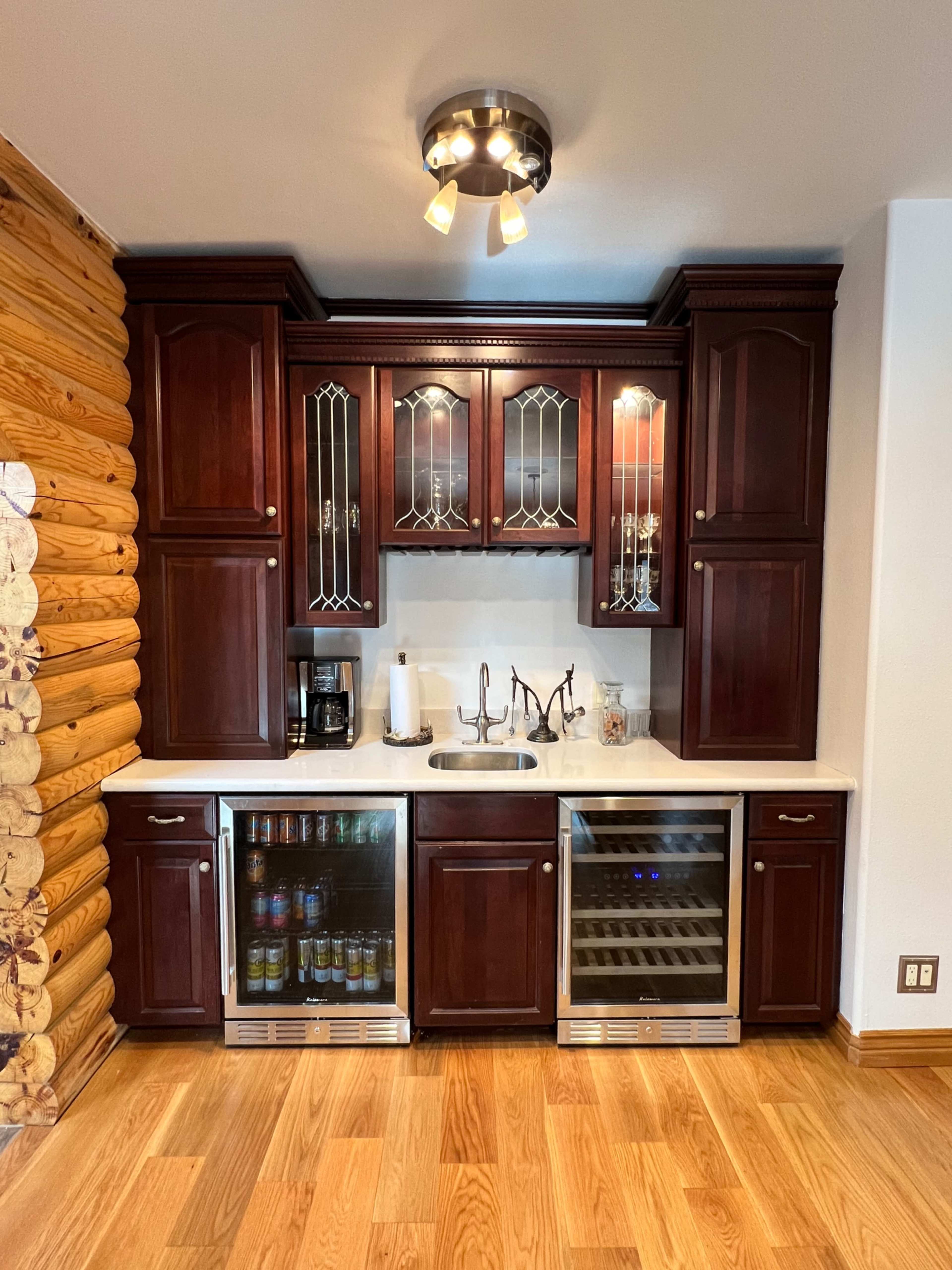 The image shows a kitchen bar area with dark wooden cabinets, a sink, a wine cooler, and a row of glassware above.