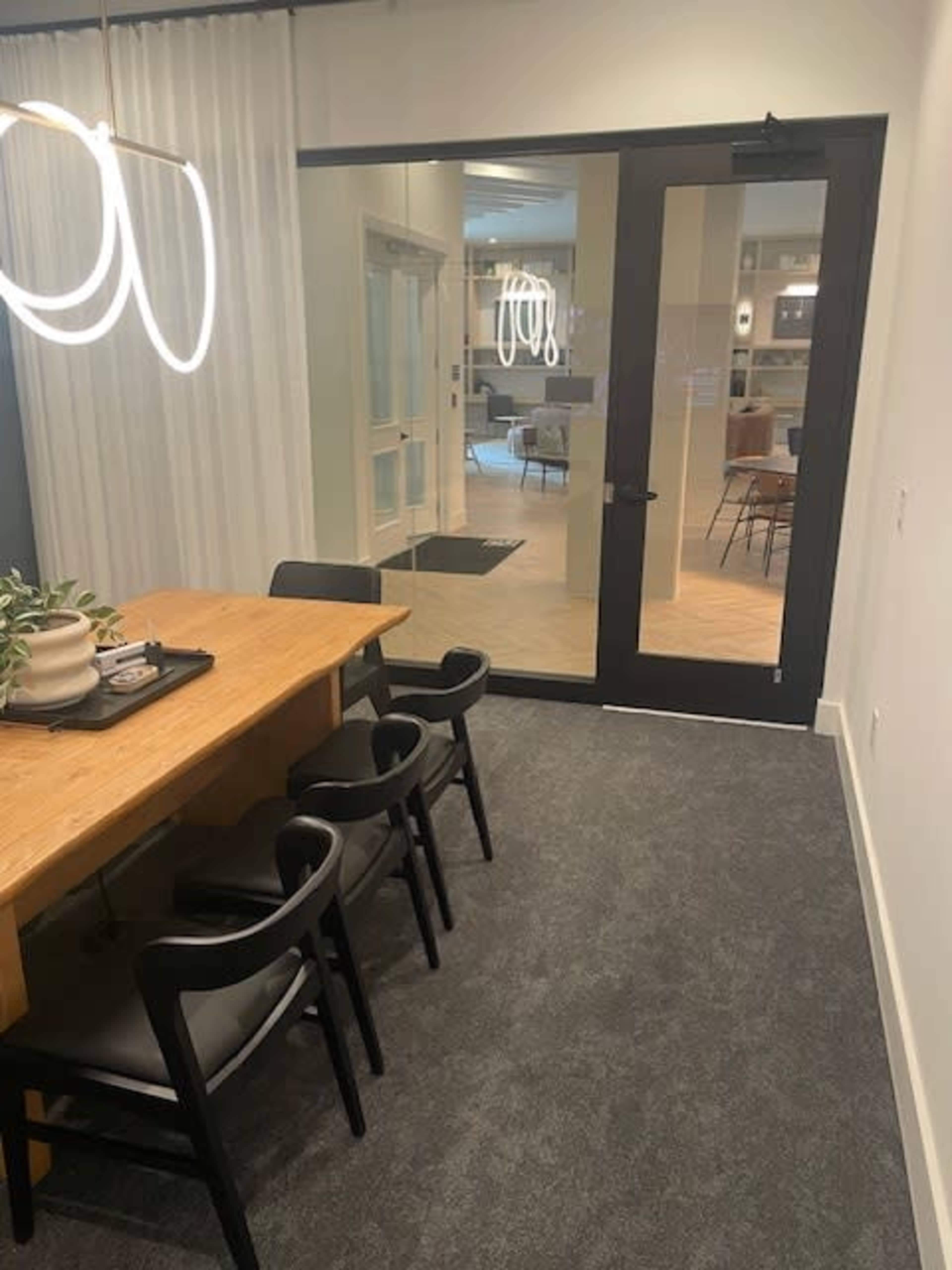 A modern conference room features a wooden table surrounded by black chairs, with a large glass door leading to a spacious area beyond.