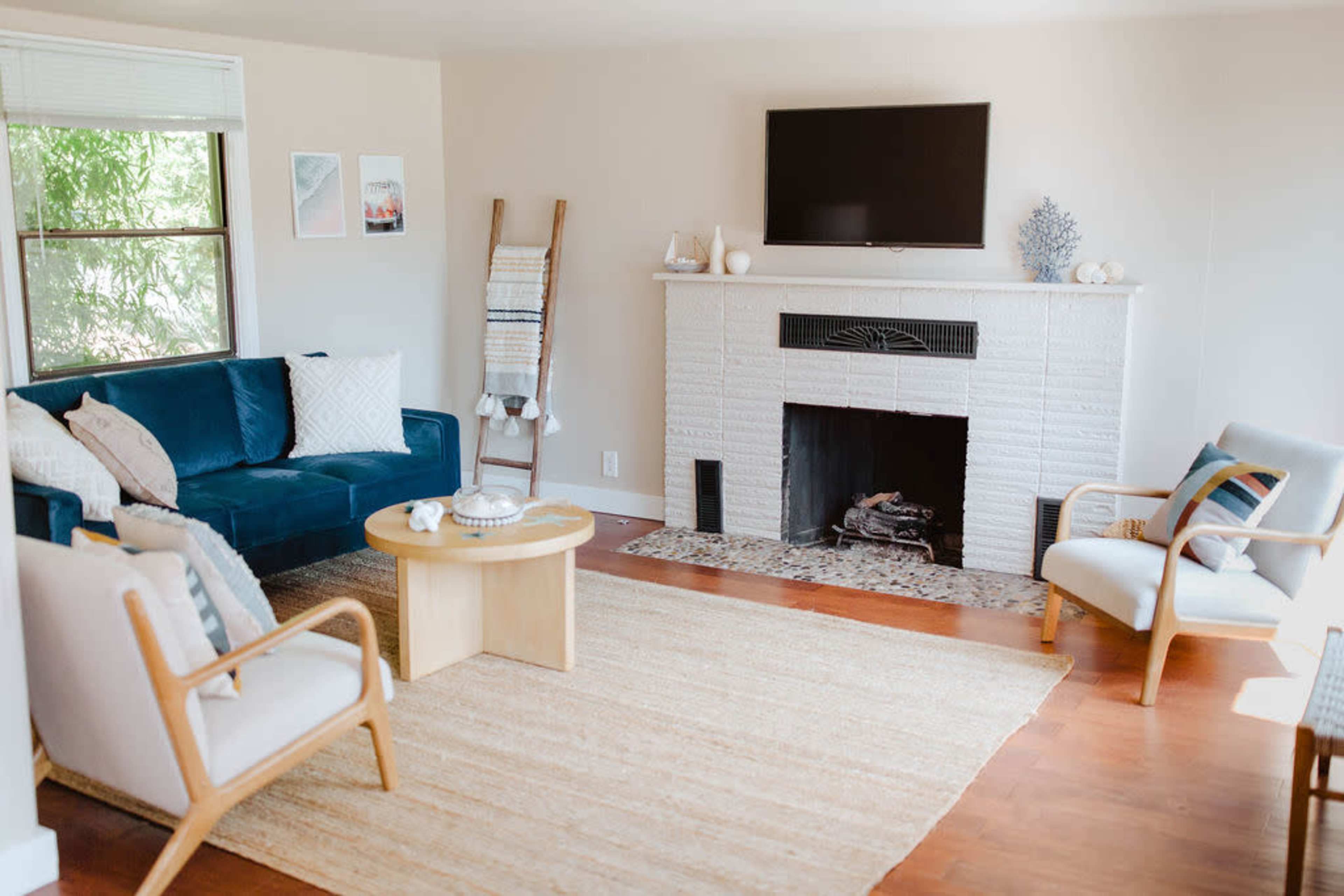 A modern living room with a blue sofa, beige chairs, a round coffee table, and a television above a white brick fireplace.