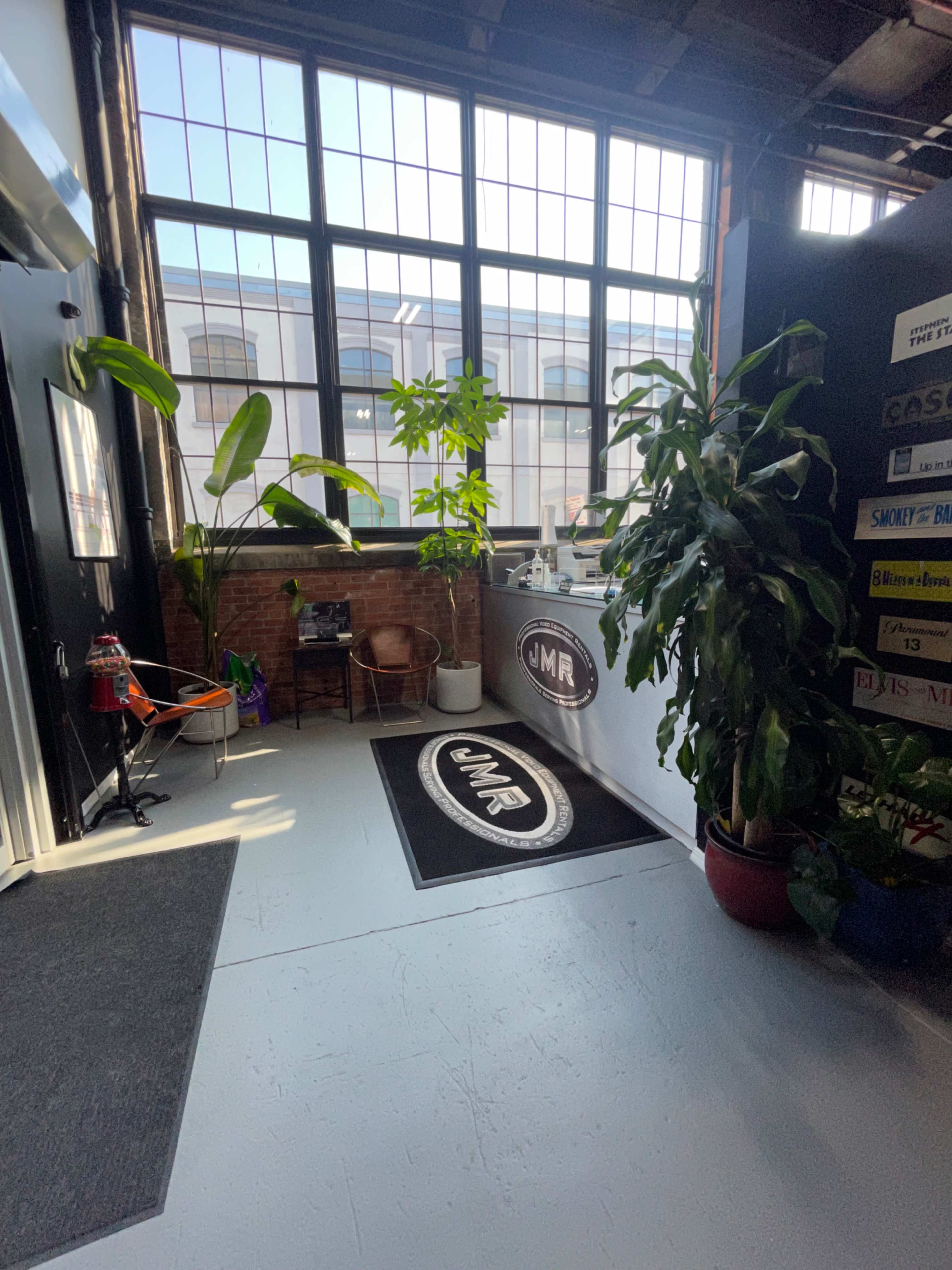 The image shows an interior space with large windows, a reception area featuring a logo mat, potted plants, and a mix of chairs and decorative elements.