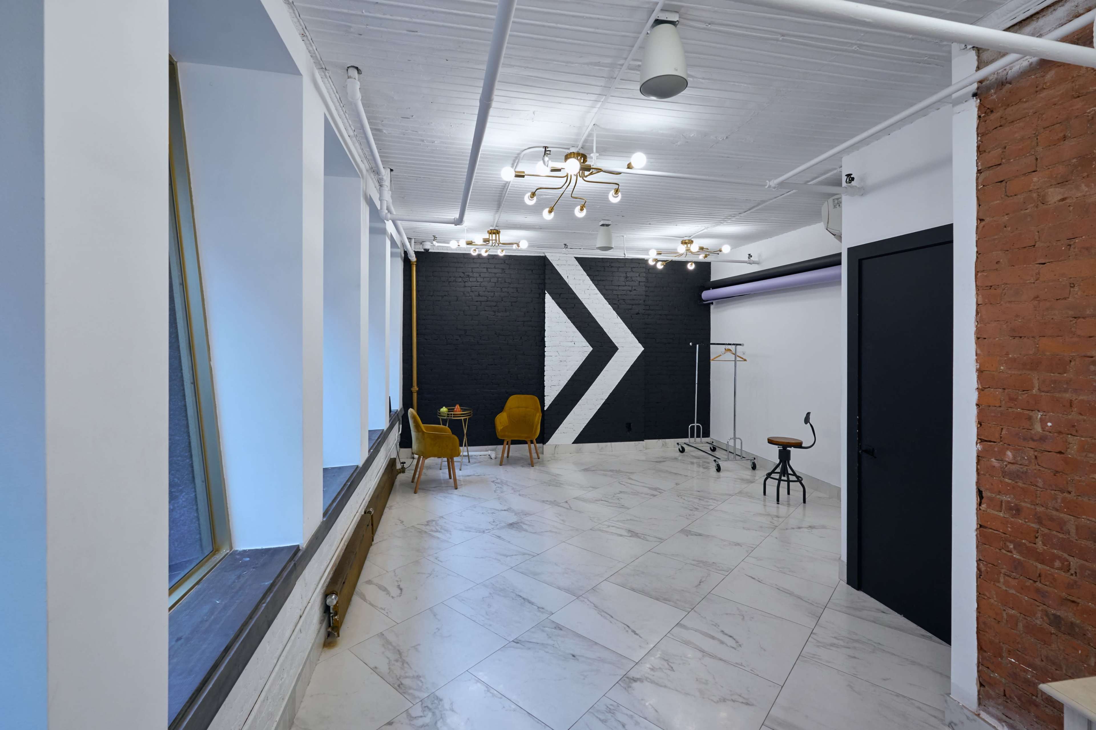 A spacious interior with a patterned black and white accent wall, marble flooring, and minimalist furniture, including two yellow chairs and a stool.