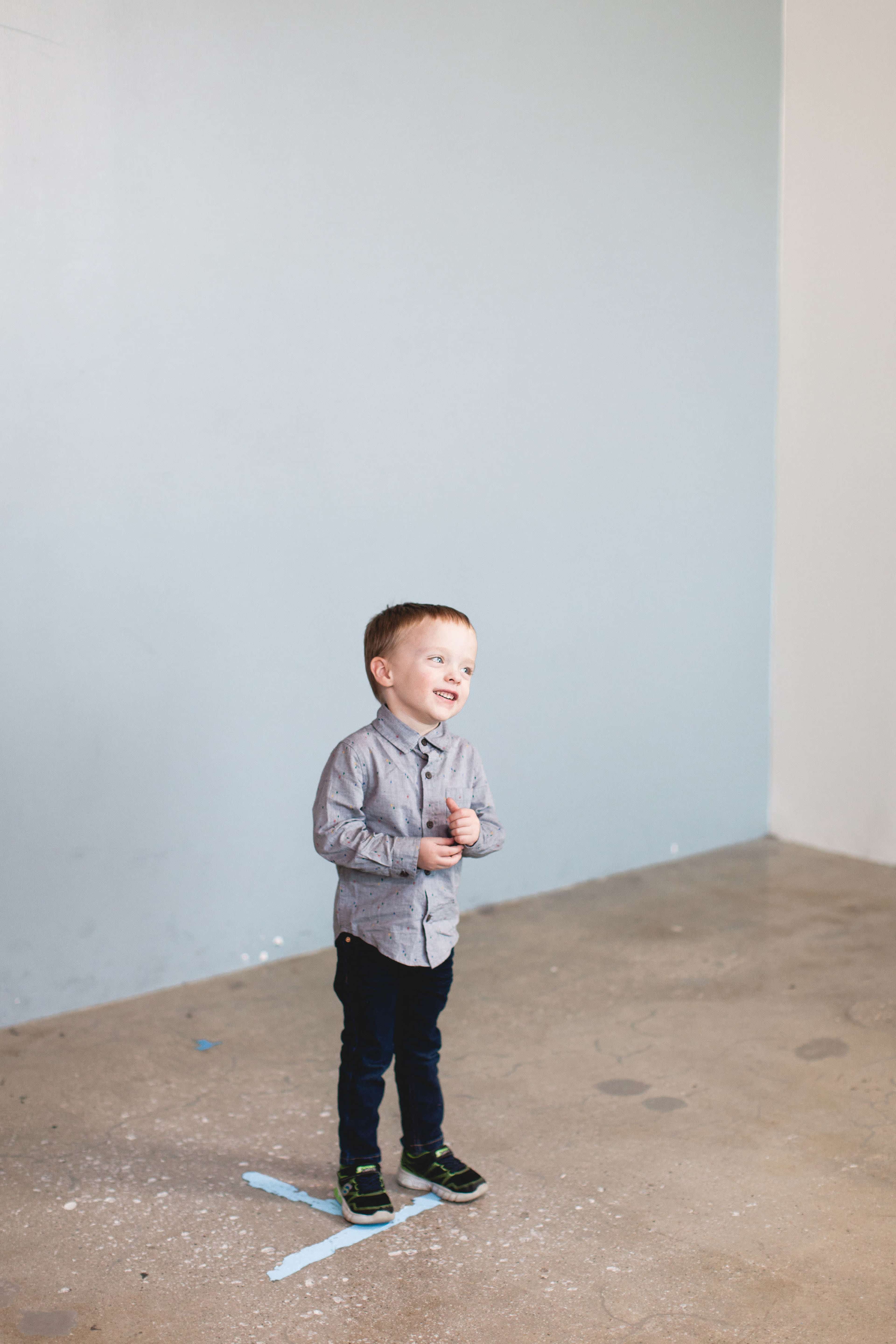 A young boy stands on a blue-marked concrete floor, smiling in front of a light-colored wall.