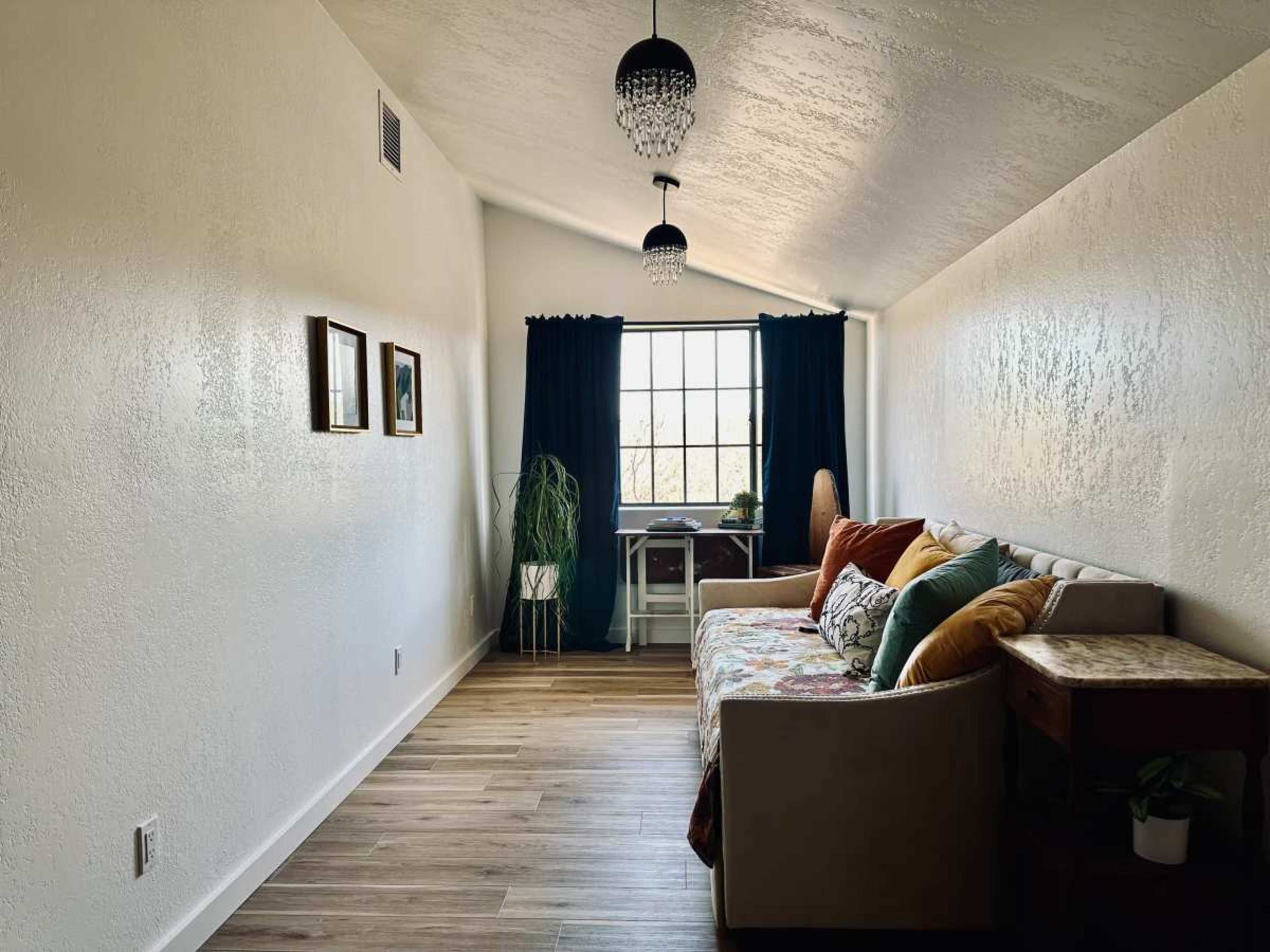 The room features a light-colored sofa adorned with decorative pillows, a wooden side table, and a window with dark curtains, all set against a wall with textured paint.