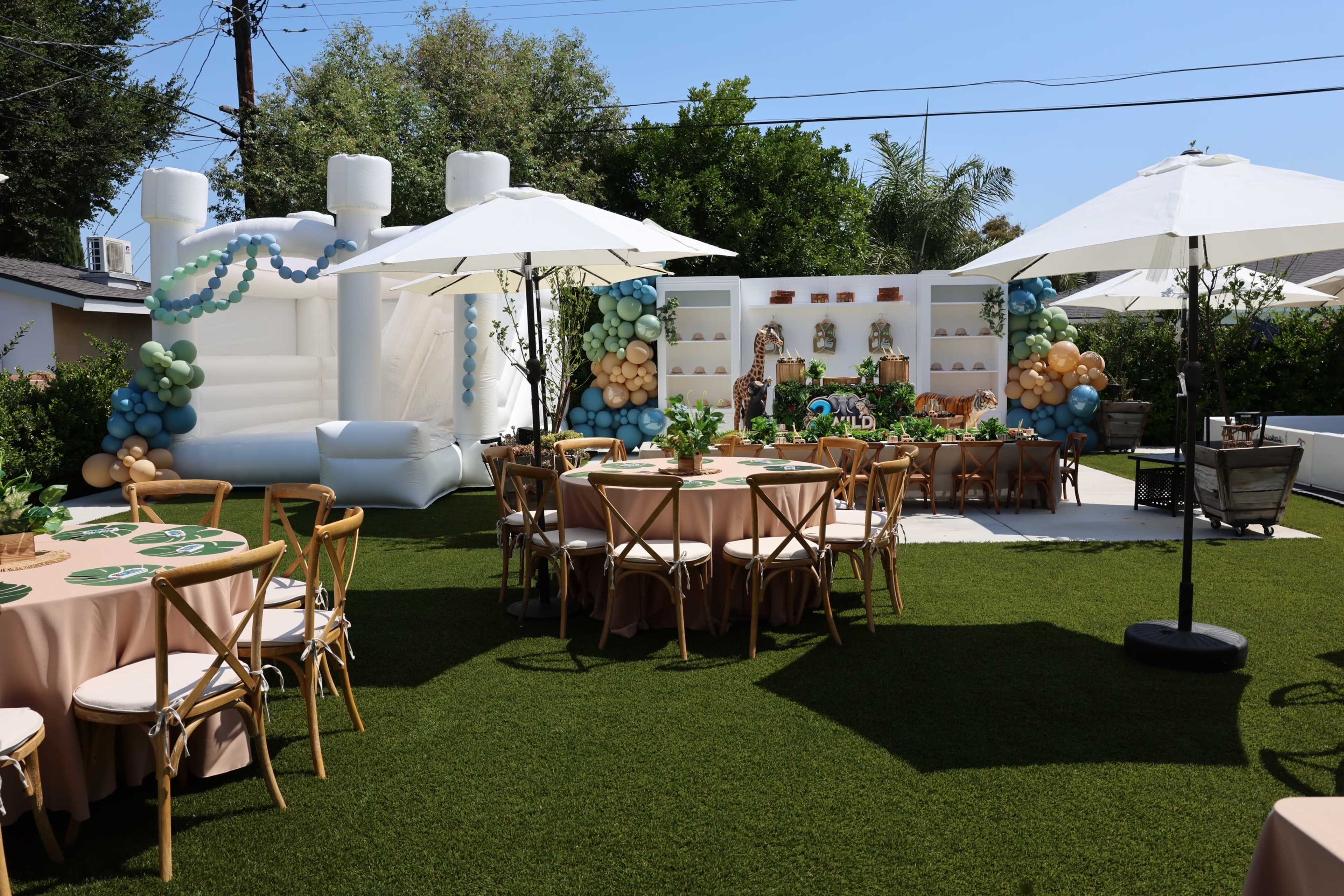 The image shows a decorated outdoor party area featuring tables with beige tablecloths, a bounce house, and colorful balloon arrangements against a backdrop of greenery.
