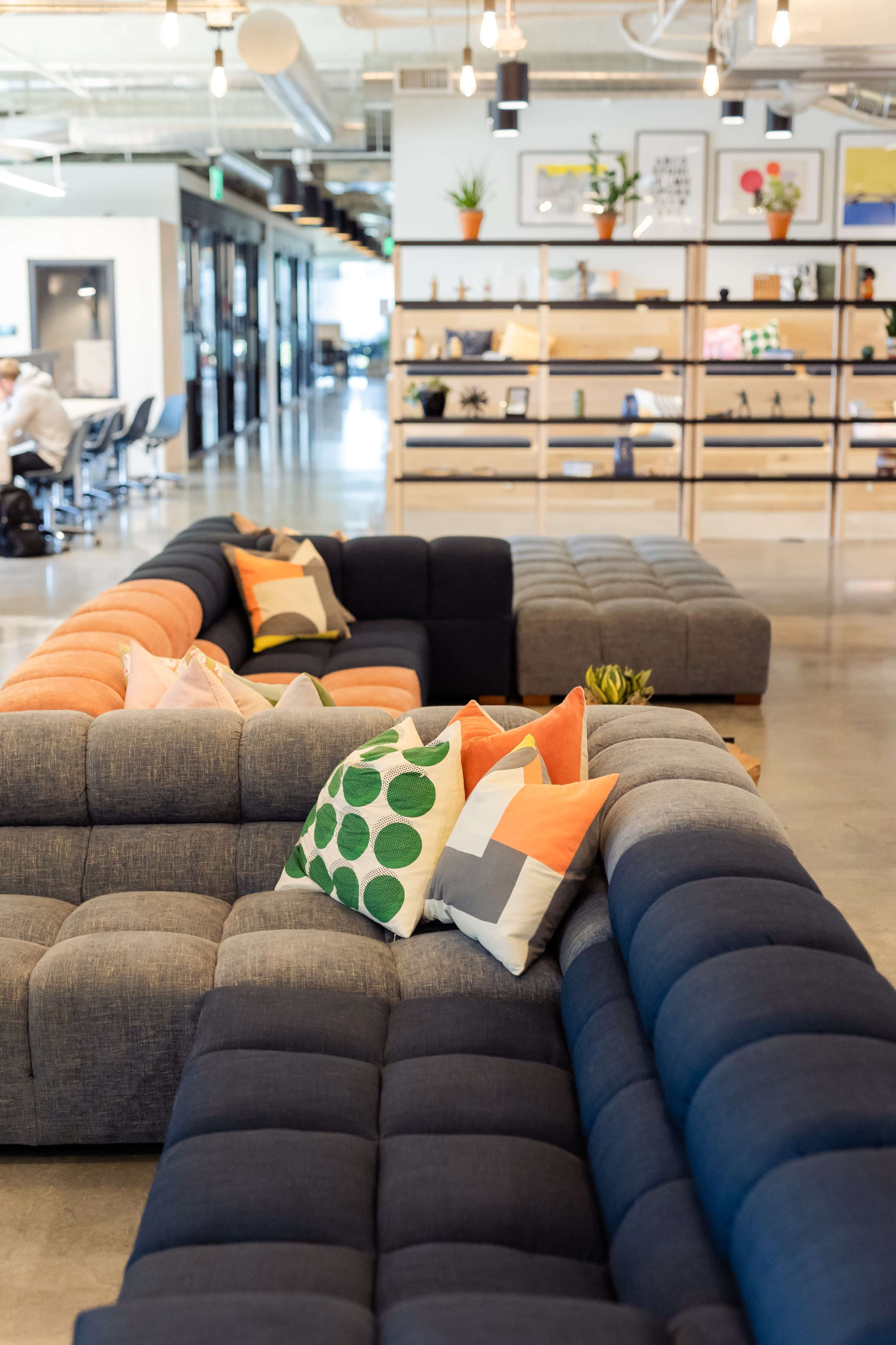 The image shows a modern office space with a large sectional sofa featuring colorful throw pillows, surrounded by a well-organized shelving unit filled with plants and decorative items.