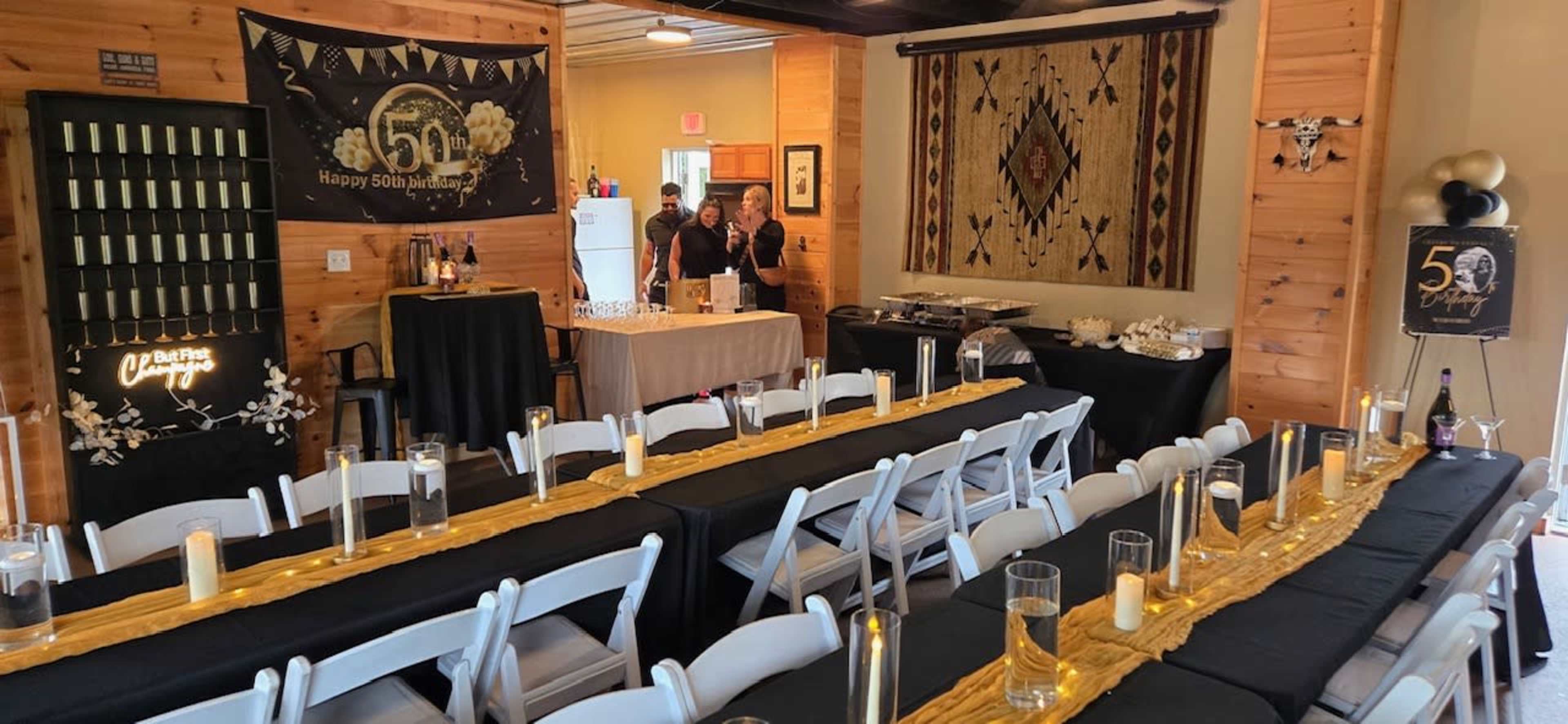 The image shows a decorated indoor venue set up for a 50th anniversary celebration, featuring tables with black tablecloths, gold accents, and a bar area in the background.