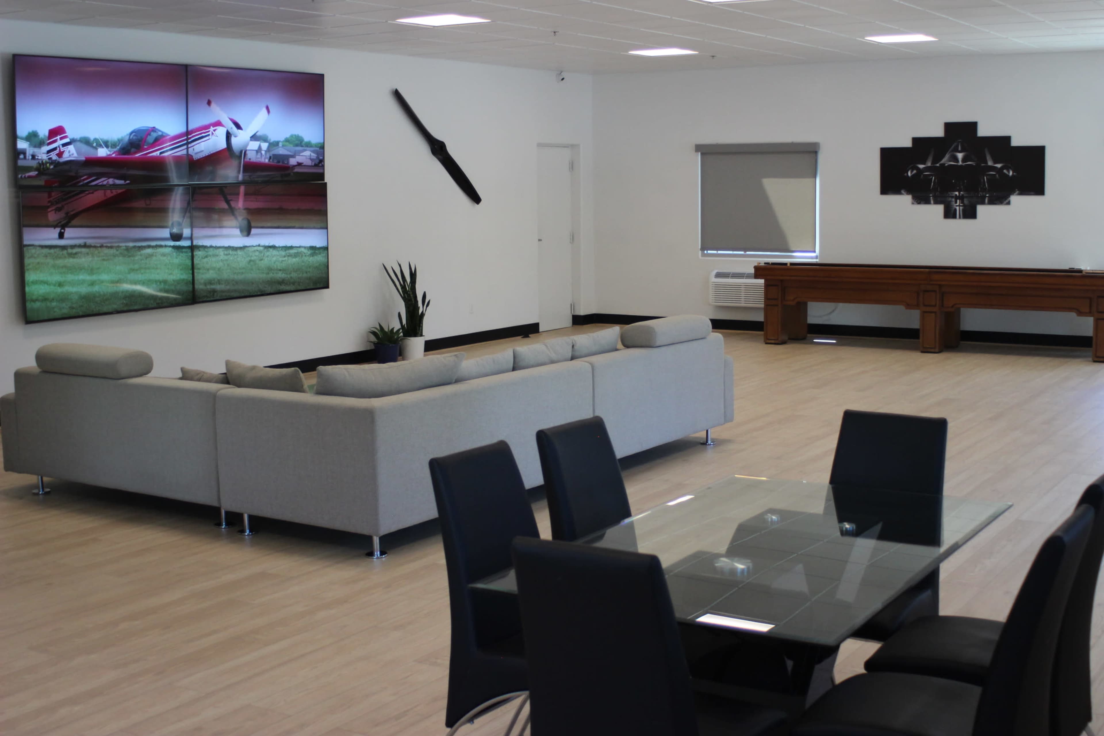 A modern interior space featuring a large sectional sofa, a glass dining table with black chairs, and a wall displaying a large digital artwork of an airplane.