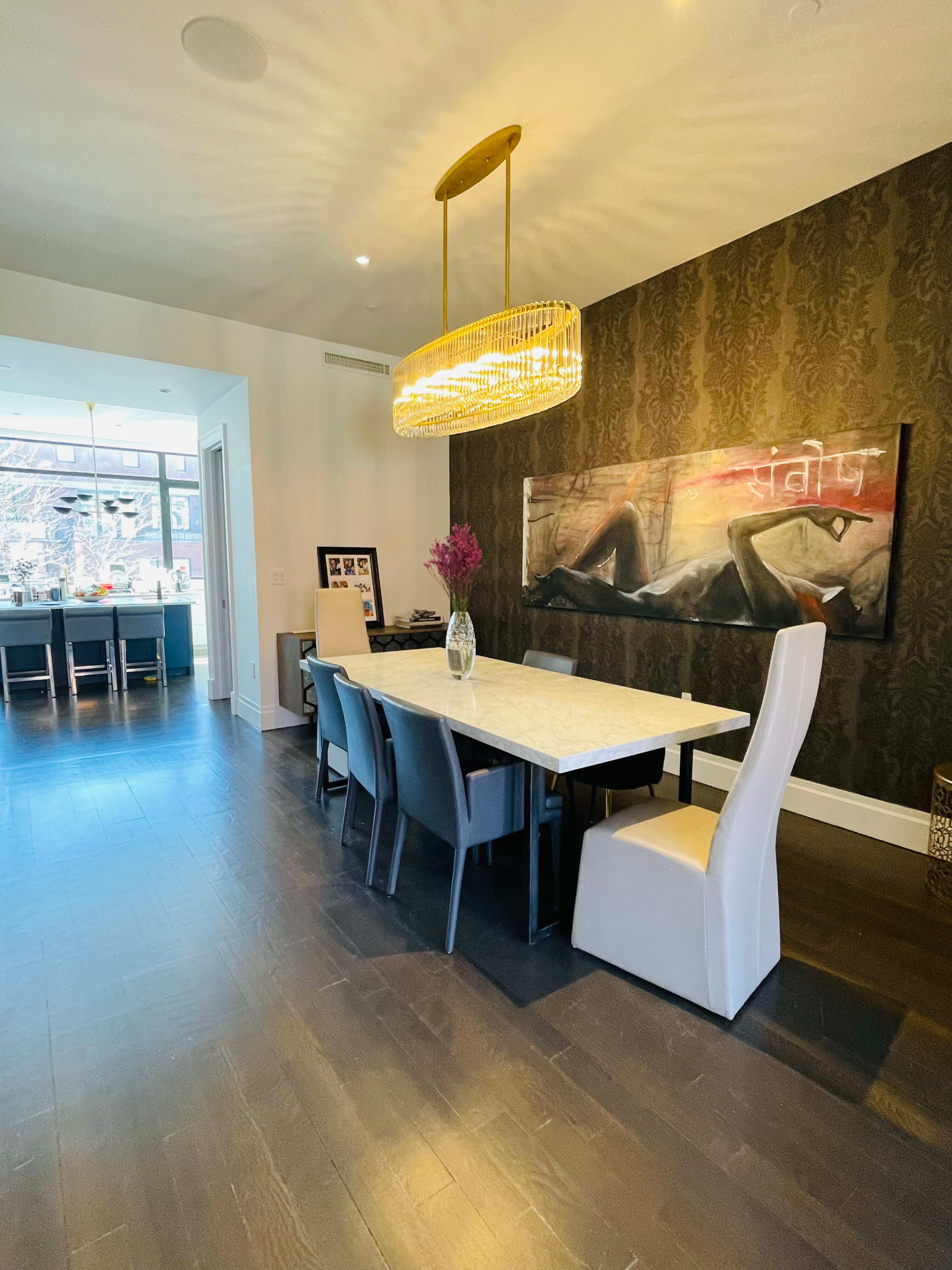 A modern dining area features a long white table surrounded by black and white chairs, with a large artwork on the wall and a pendant light overhead.