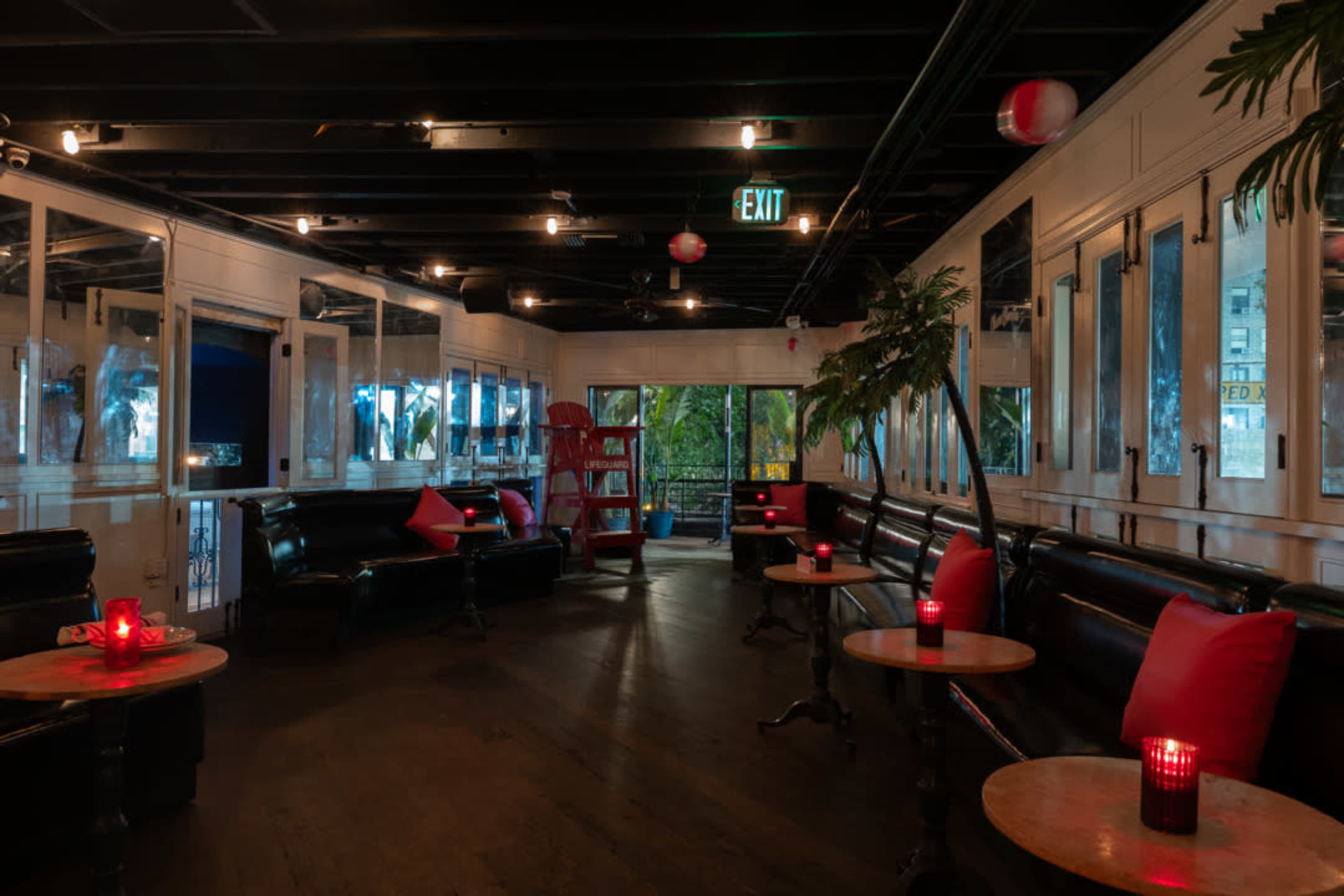 The image shows a dimly lit lounge with black sofas, red cushion accents, wooden tables, and a tropical plant, alongside a bar area visible through large windows.
