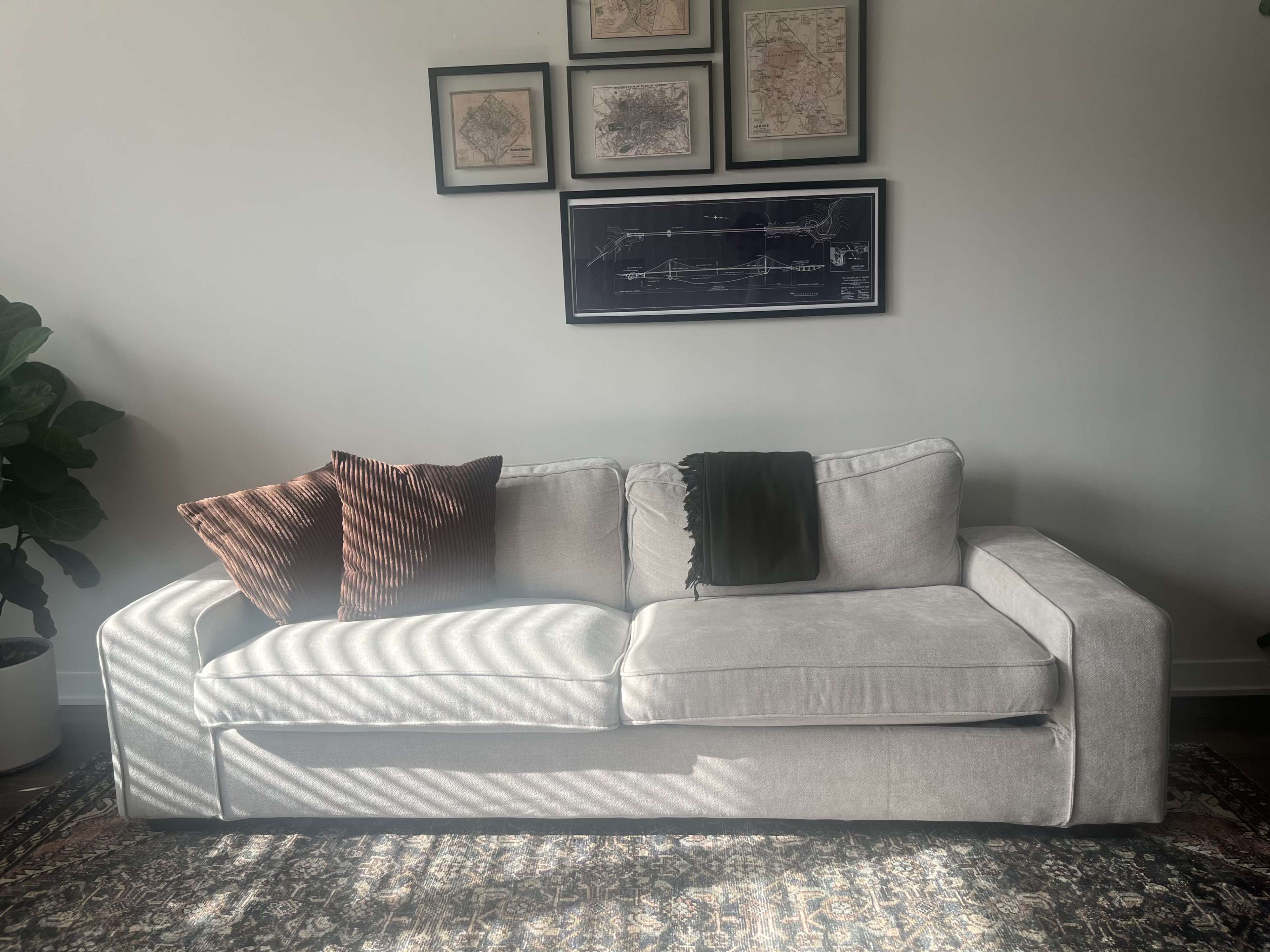 A light-colored sofa with two decorative pillows and a throw blanket is positioned against a wall adorned with framed maps.