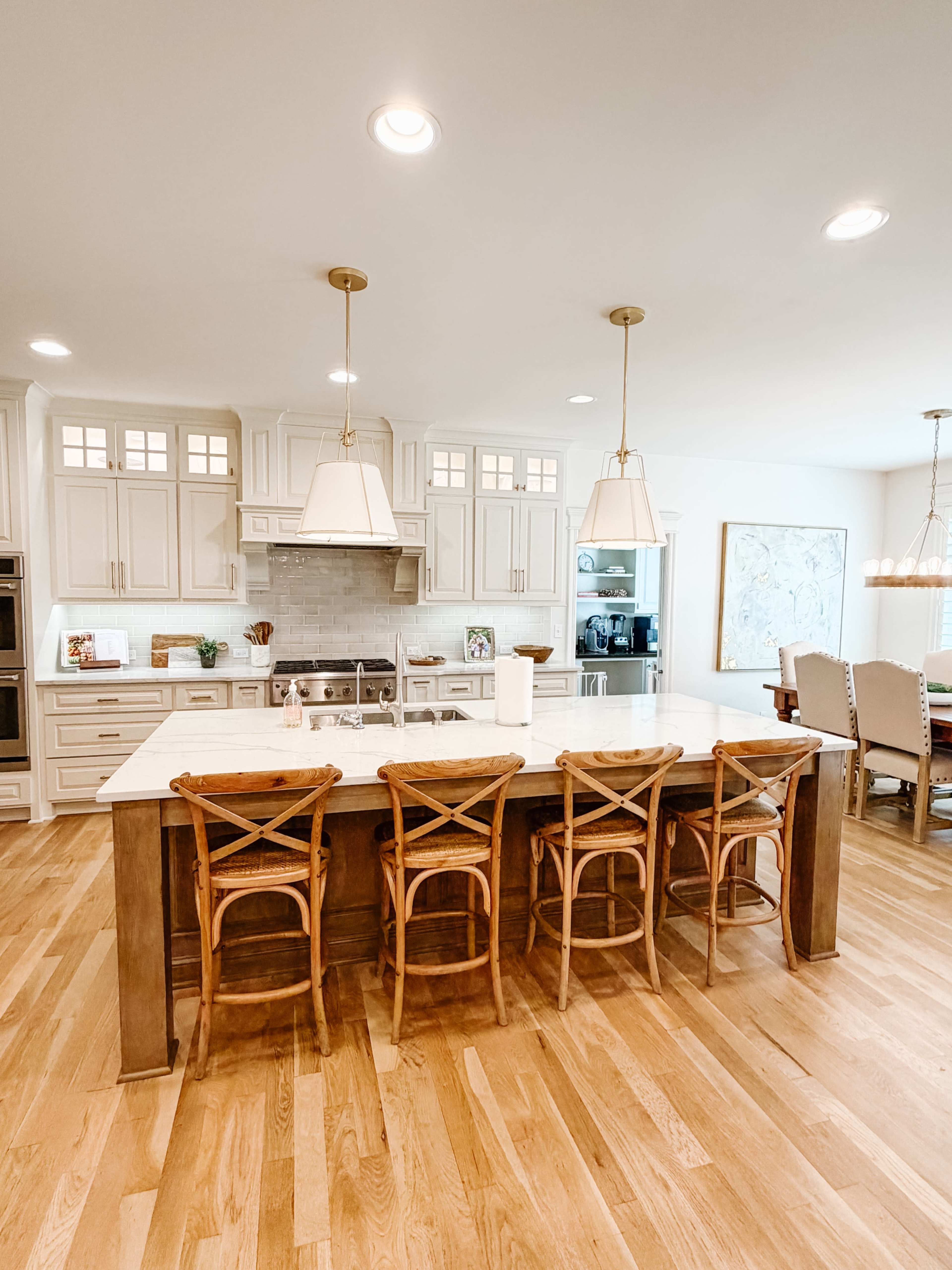 The image shows a spacious kitchen with light-colored cabinets, a large island with wooden stools, and a dining area featuring a wooden table.