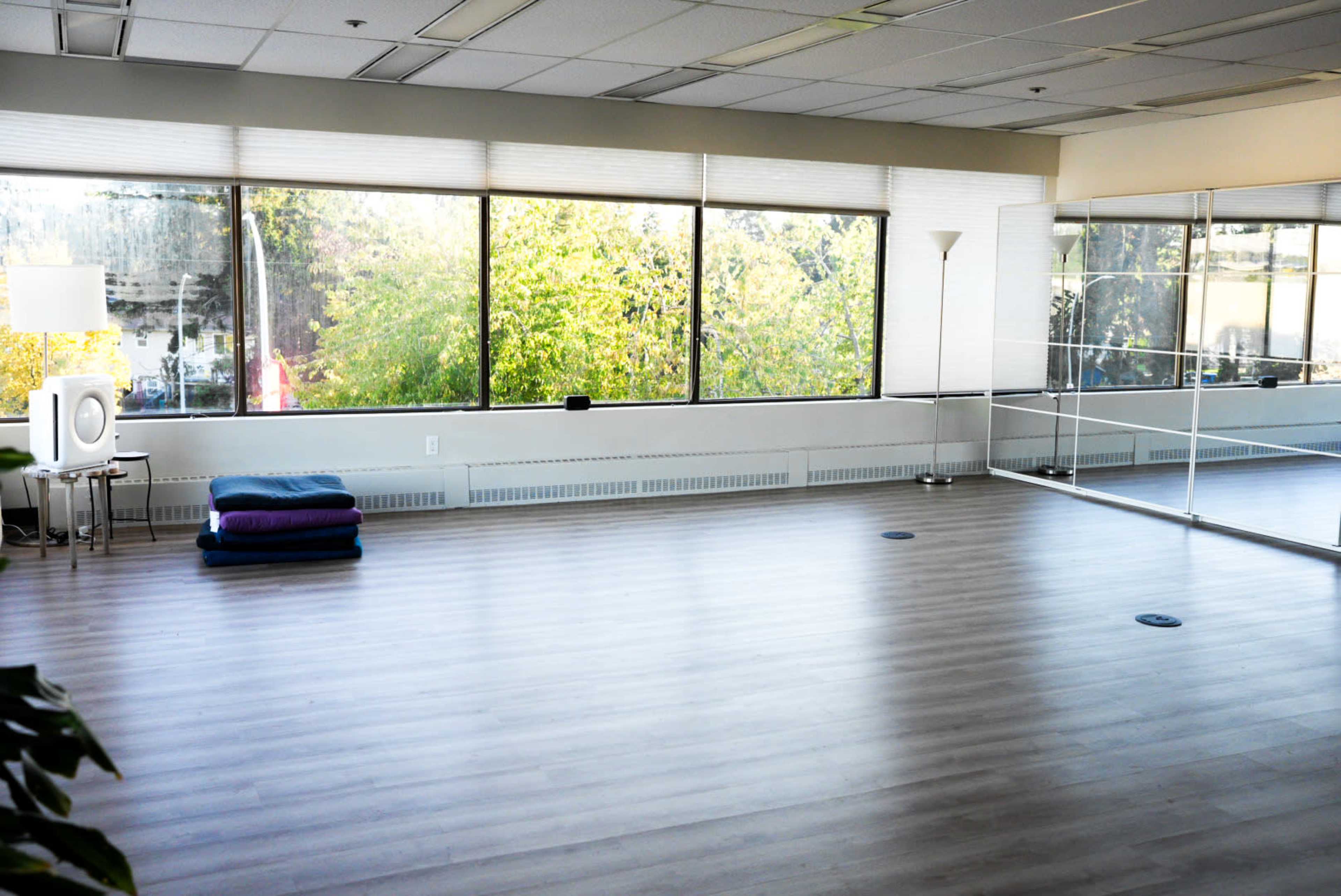 A spacious, well-lit room with large windows, hardwood floors, and a mirror wall, featuring a stack of blue blankets in one corner.