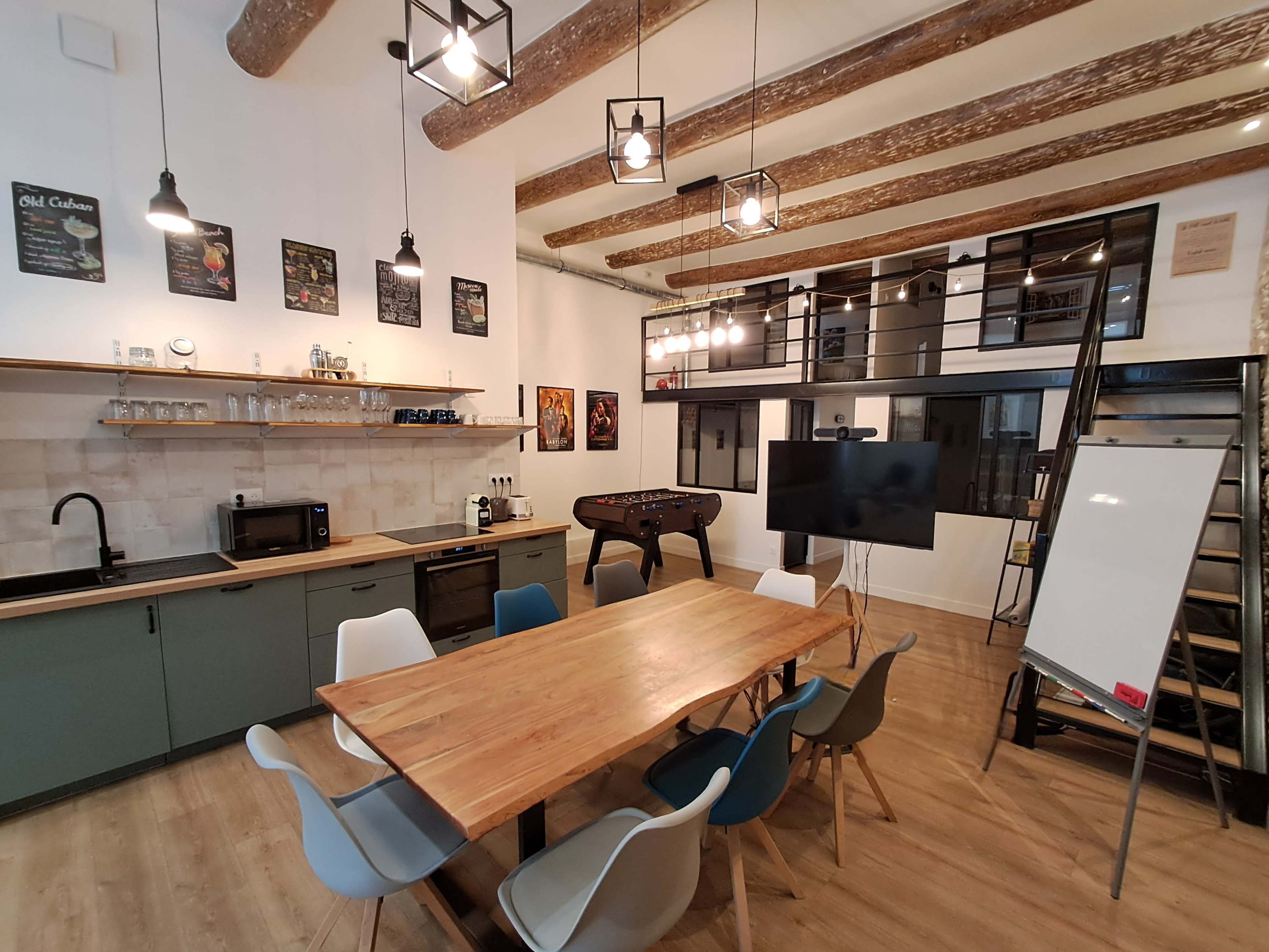 The image shows a modern meeting room with a wooden table surrounded by chairs, a kitchenette along one wall, a foosball table, and a staircase leading to an upper level.
