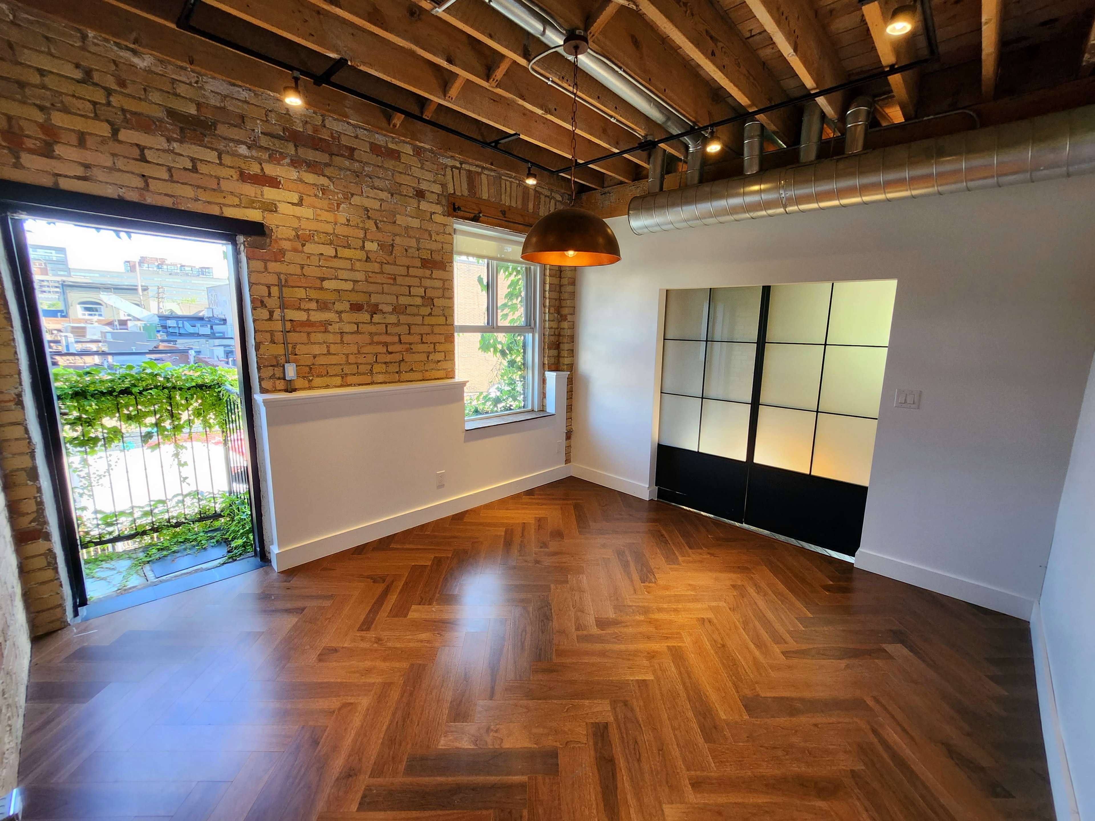 A room with exposed brick walls, wooden flooring in a herringbone pattern, a large window facing a balcony, and a decorative black partition.