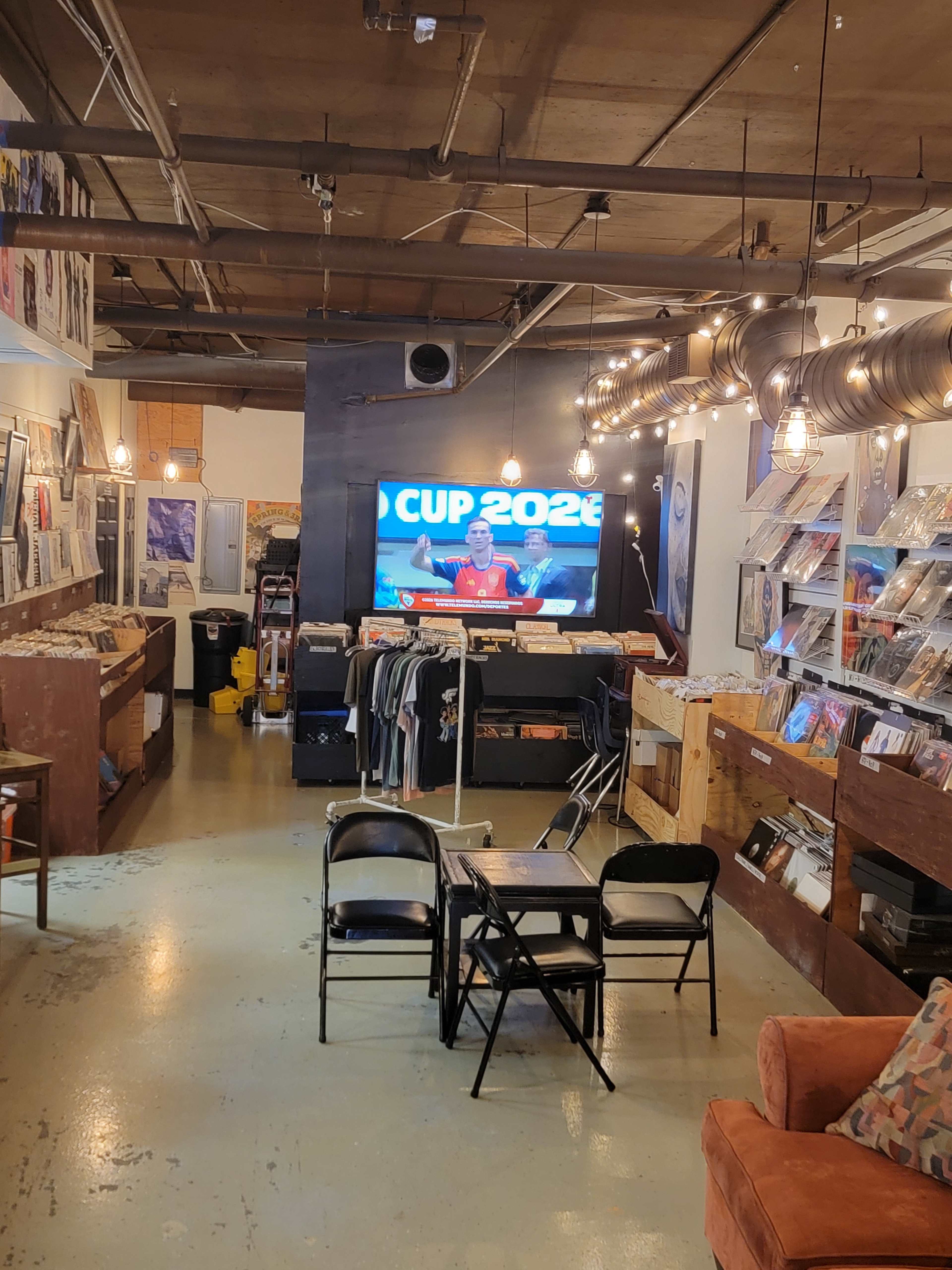 The image shows a vinyl record store interior featuring shelves of records, chairs arranged around a small table, and a large screen displaying a sports event titled "CUP 2026" on the wall.