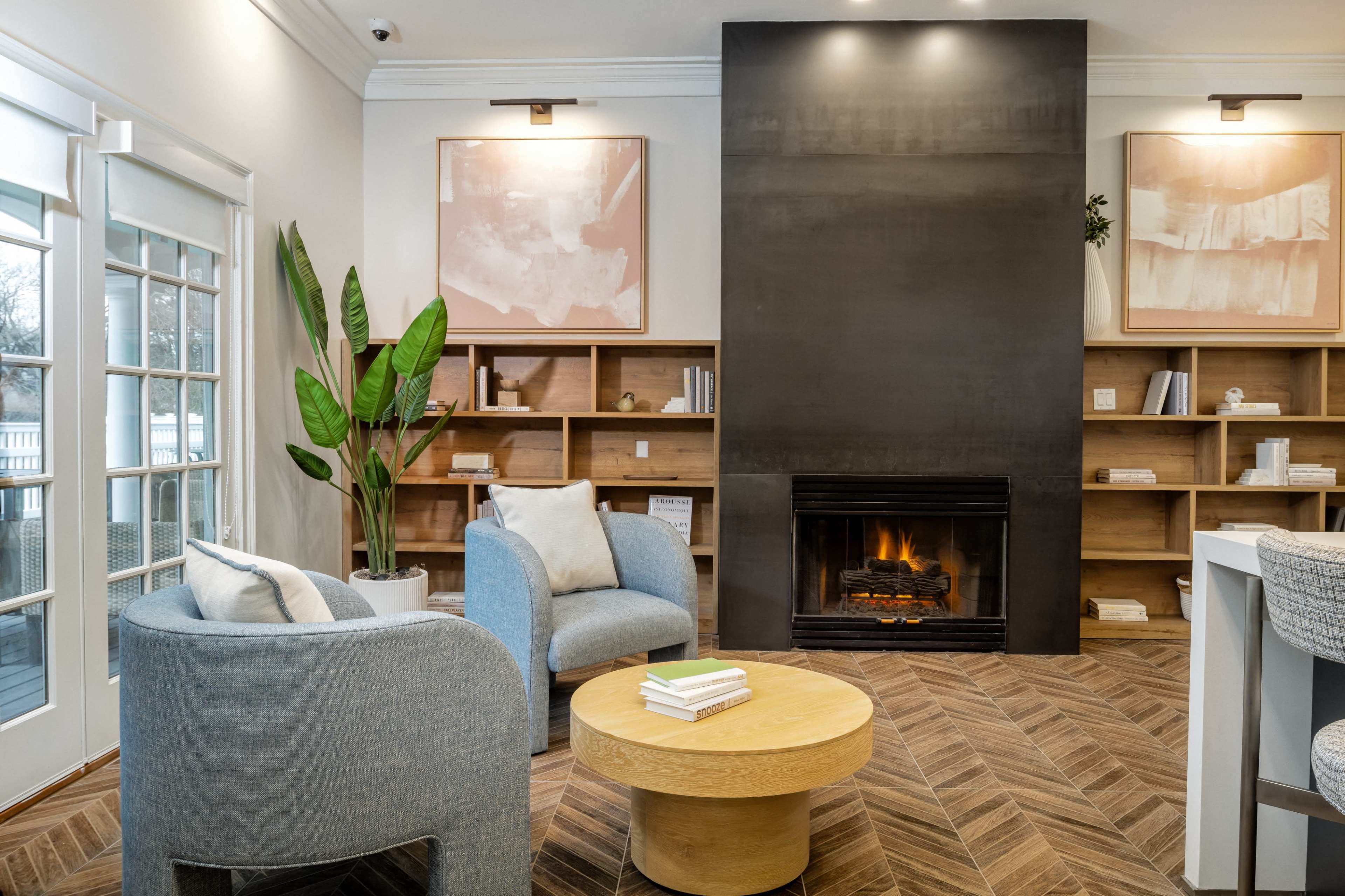 A cozy interior space featuring two light blue armchairs, a round wooden coffee table, and a modern fireplace surrounded by wooden shelving.