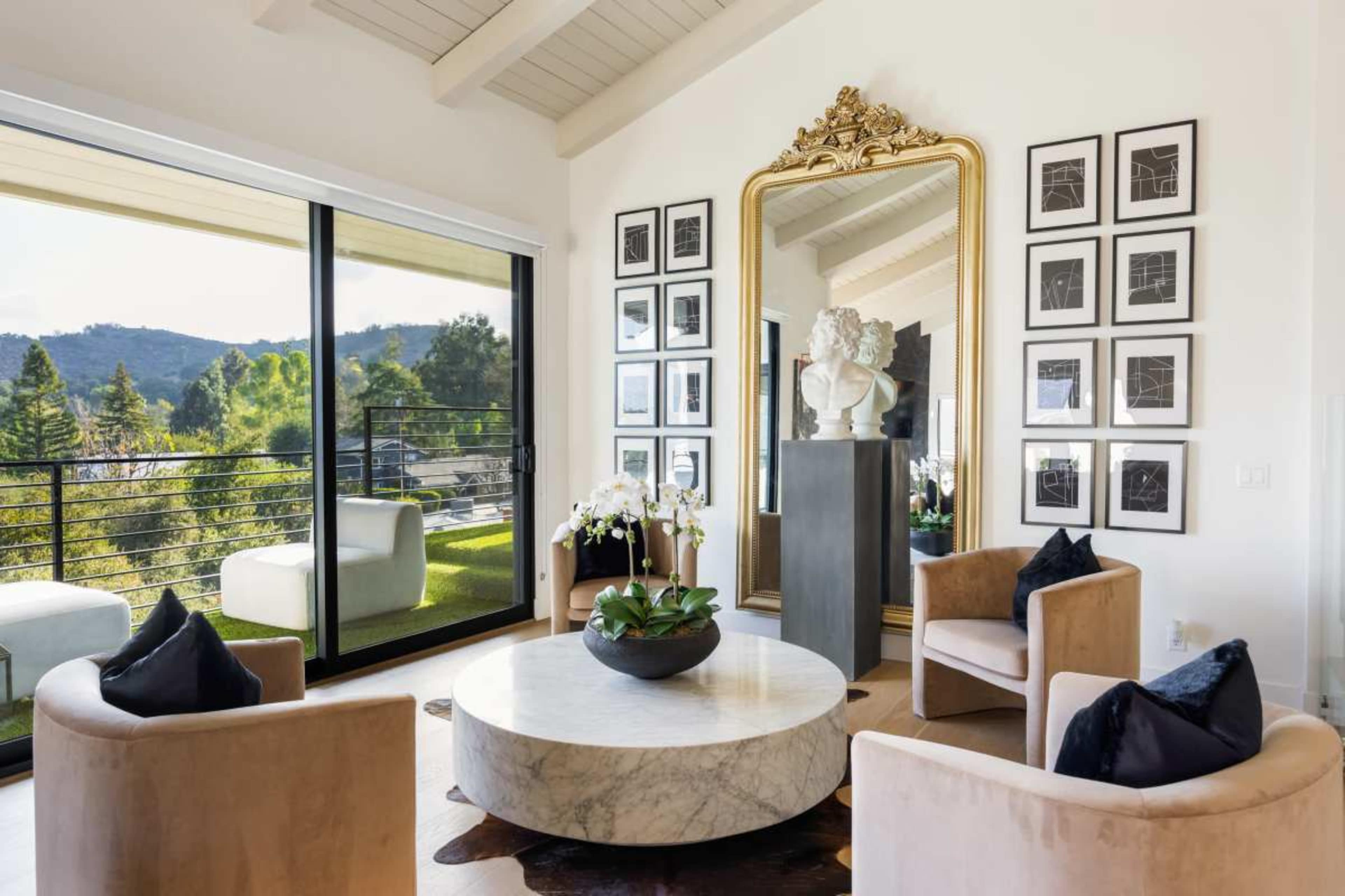 The image depicts a modern living room with two upholstered chairs, a round marble coffee table, framed artwork on the walls, and large glass doors opening to a scenic view.