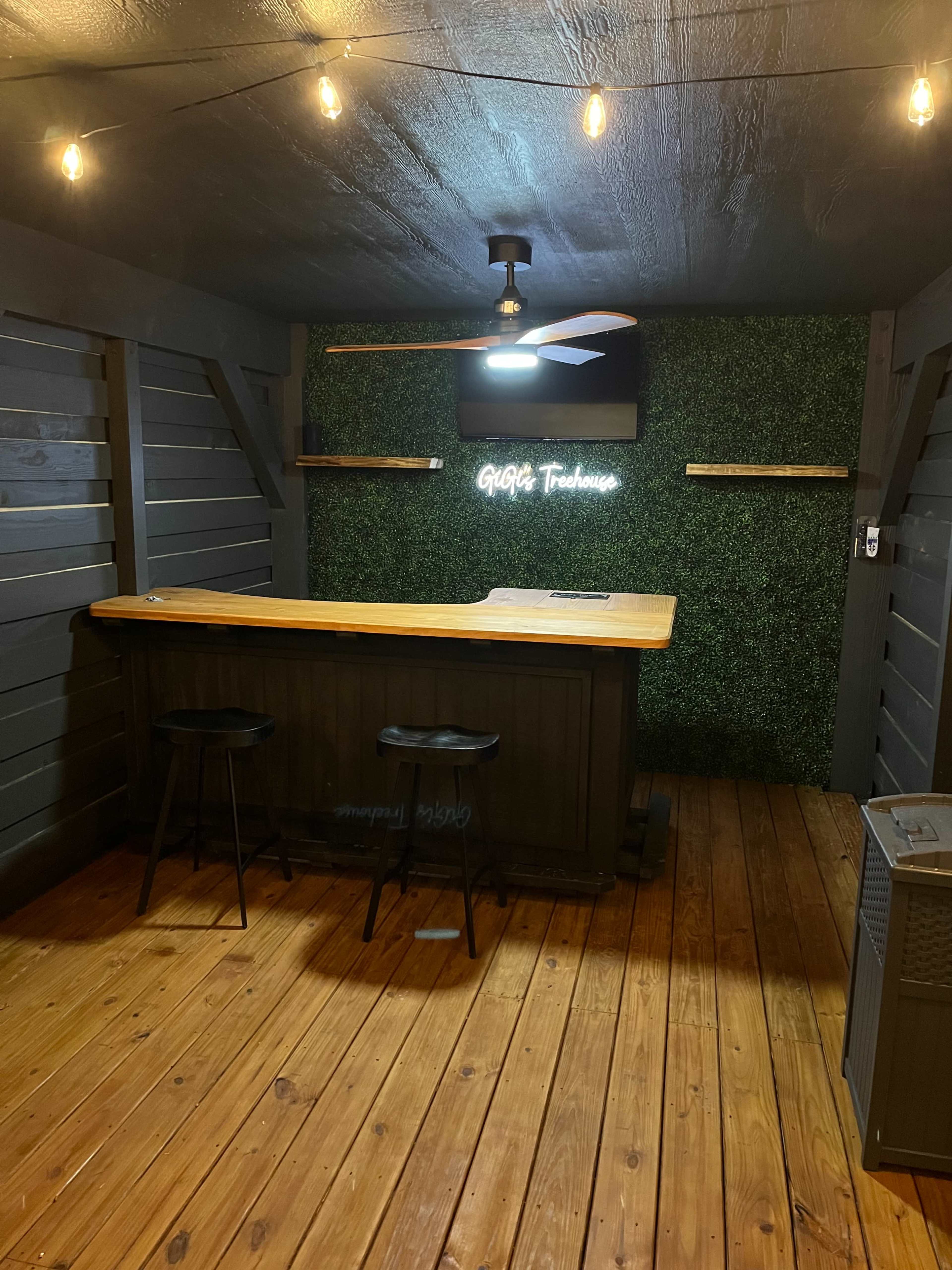 The image shows a rustic indoor space featuring a wooden bar with two black stools, a ceiling fan, and a neon sign against a green wall, illuminated by string lights.