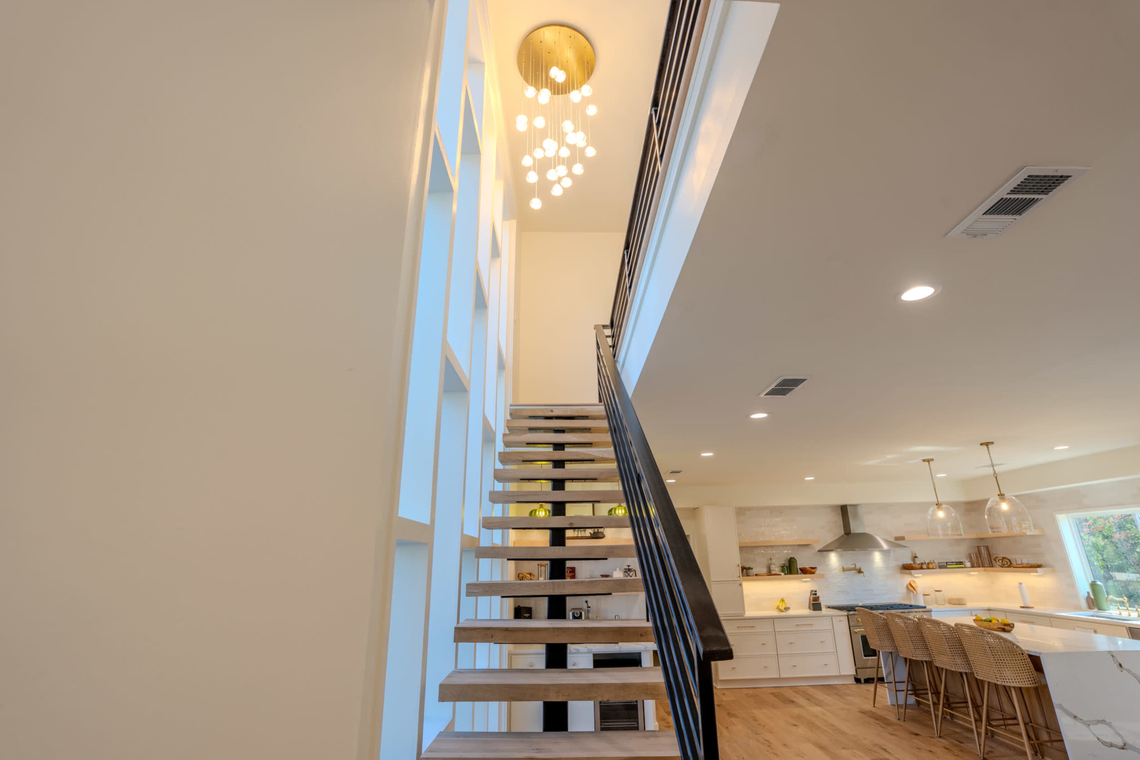 The image shows a modern staircase leading to an upper level, with a pendant light above and a view of a kitchen in the background.
