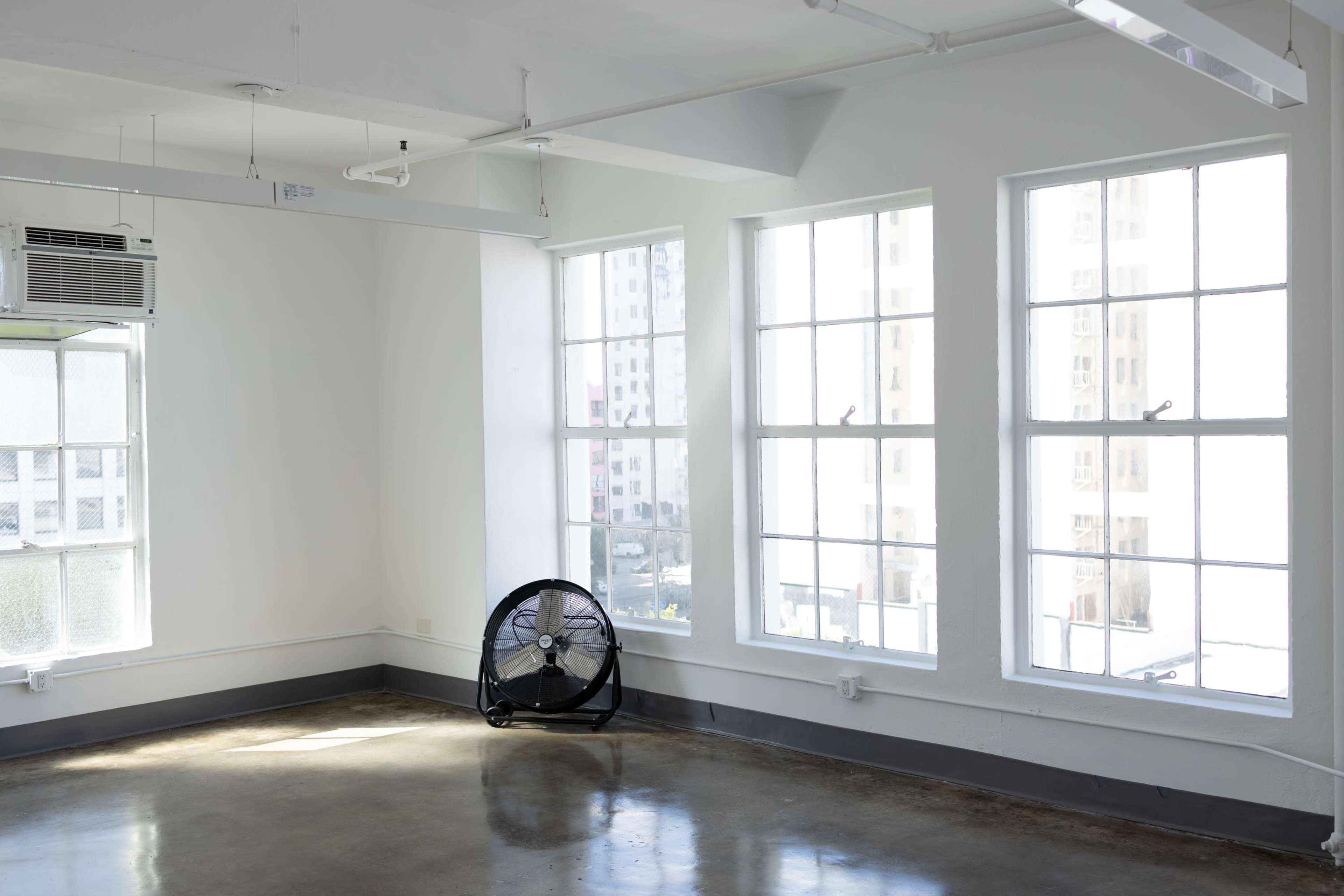 A large, empty room with concrete floors and multiple windows allowing natural light, featuring a standing fan in one corner.
