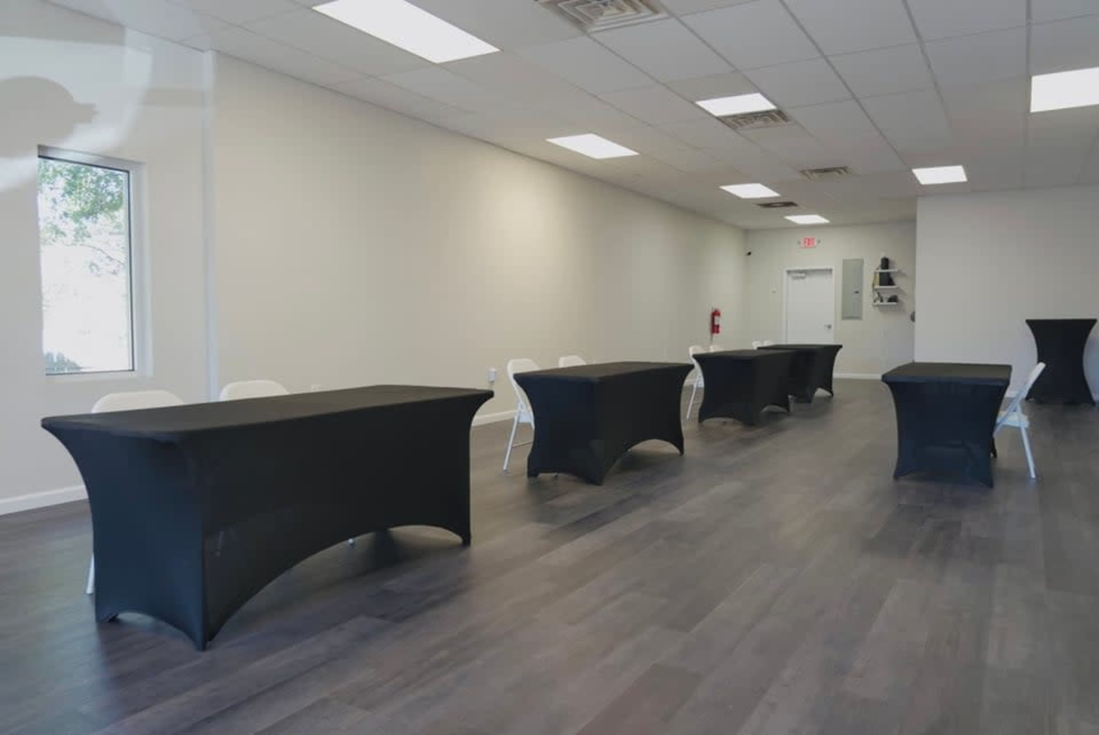 The image shows a spacious, well-lit room with several black tables arranged in a row and white chairs placed at each table.