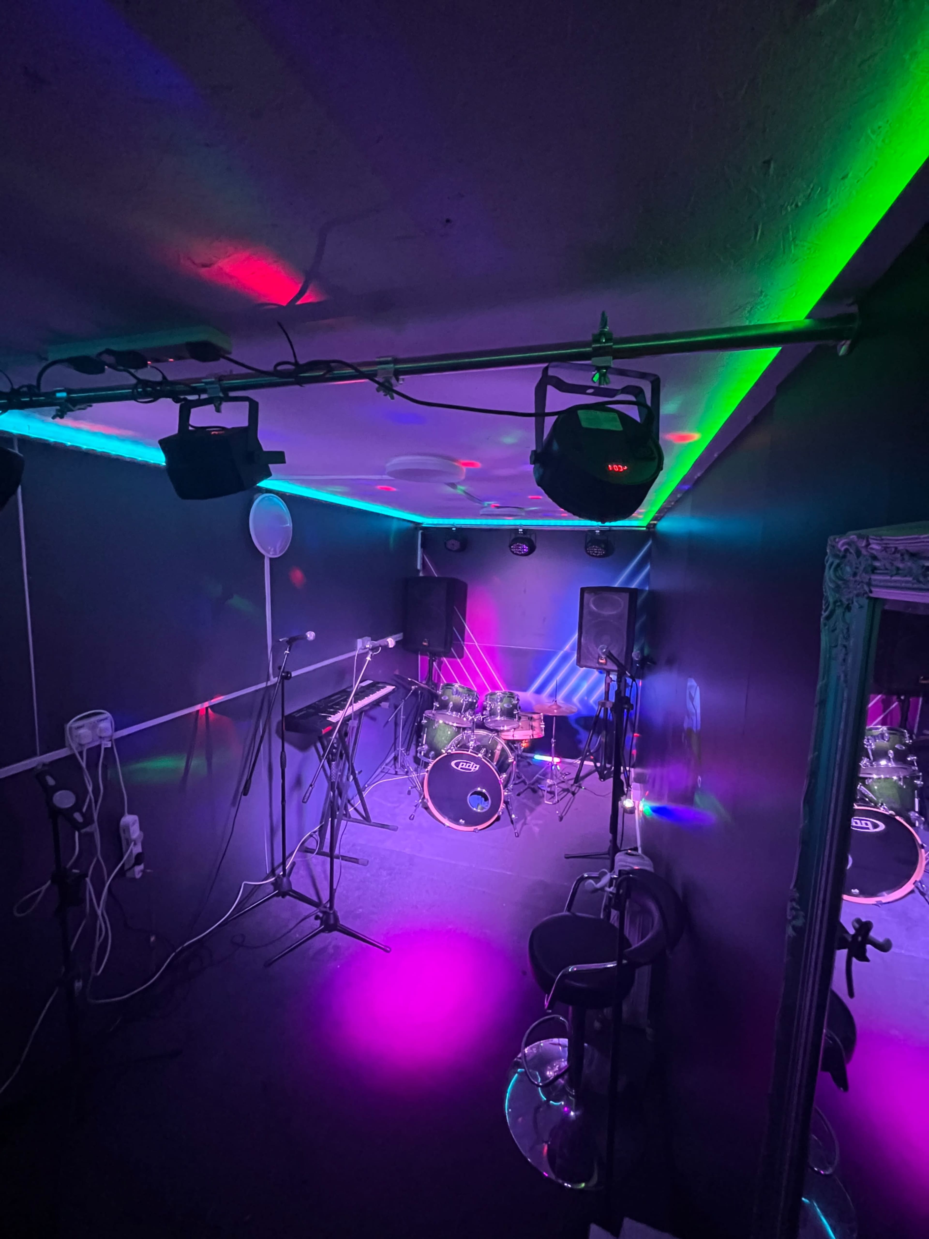The image shows a small music rehearsal studio with a drum set, microphones, and colorful lights illuminating the space.